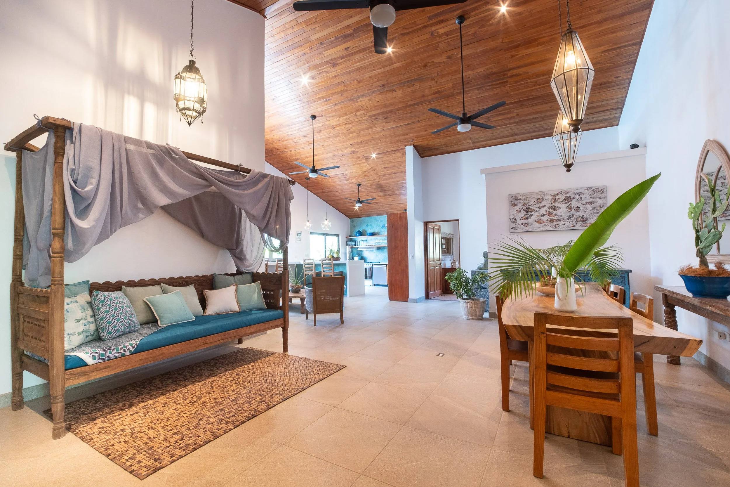 Open-concept living room and dining area with wooden ceiling, ceiling fans, chandeliers, plants, and modern decor.