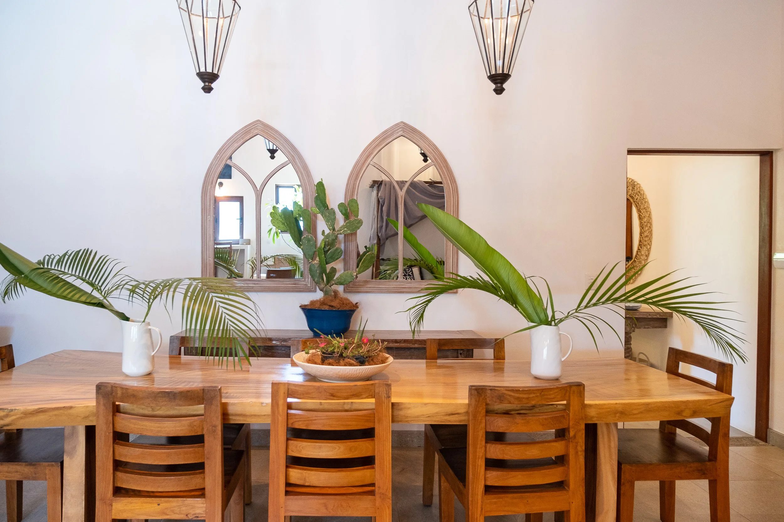 A wooden dining table with six matching chairs, decorated with large potted plants in white vases and a central plant in a bowl. Two arched mirrors hang on the white wall behind, and there are two pendant lights above. A small mirror with a woven fra