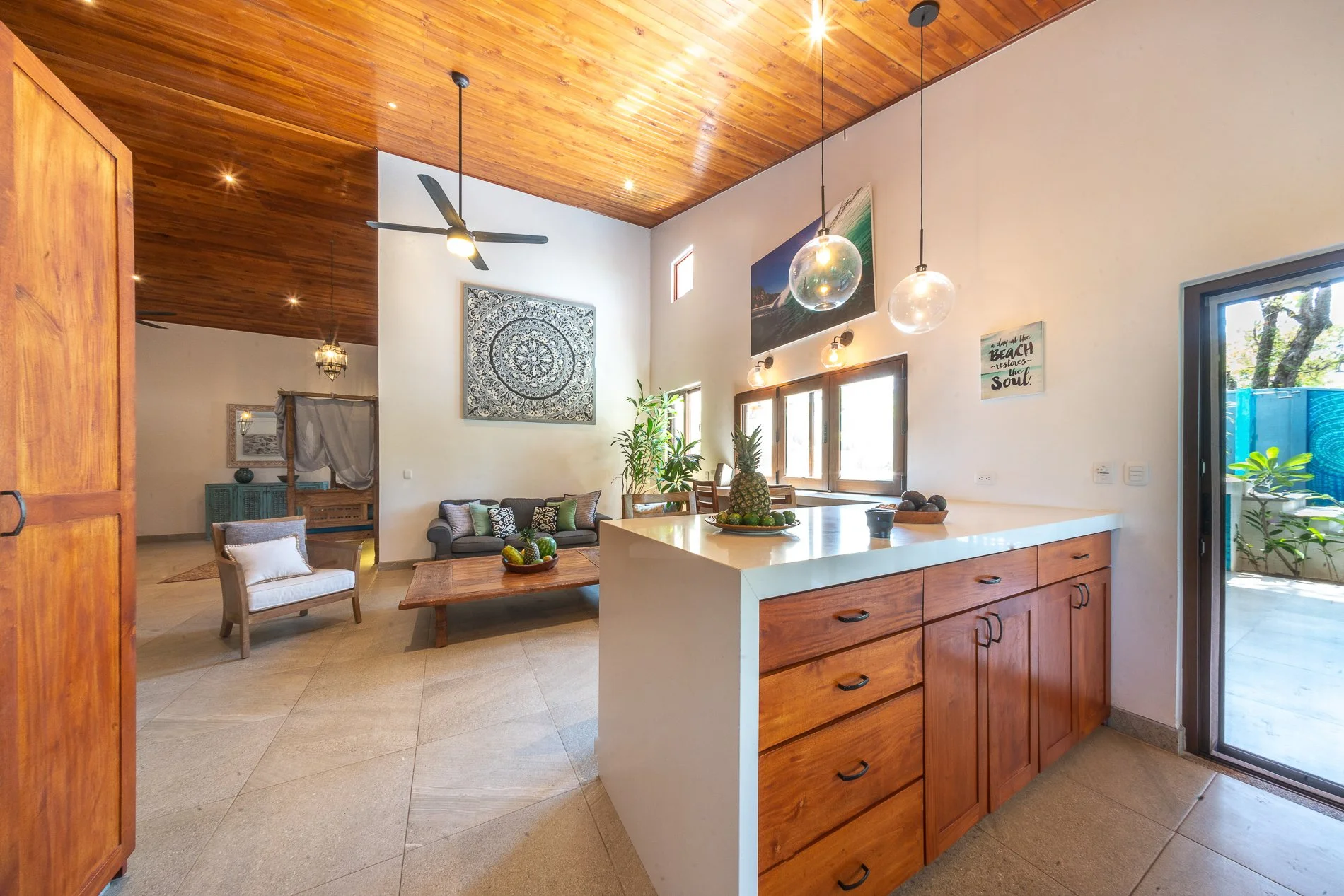 Open-concept living room with wooden ceiling, beige floor tiles, sofa, armchair, mid-century coffee table, wall art, houseplants, and a kitchen island with fruit bowls, overlooking outdoor patio.