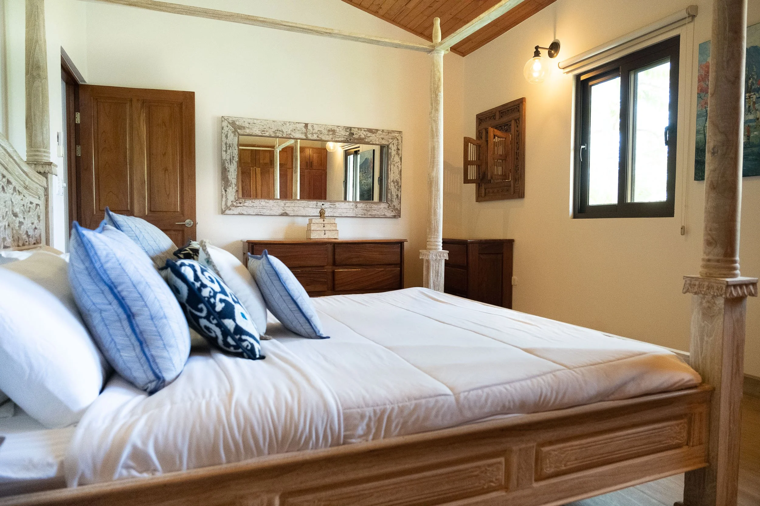 A bedroom with a wooden four-poster bed, decorated with blue and white pillows, a mirror on the wall, a wooden dresser, and a window with black frame, in a cozy rustic style.