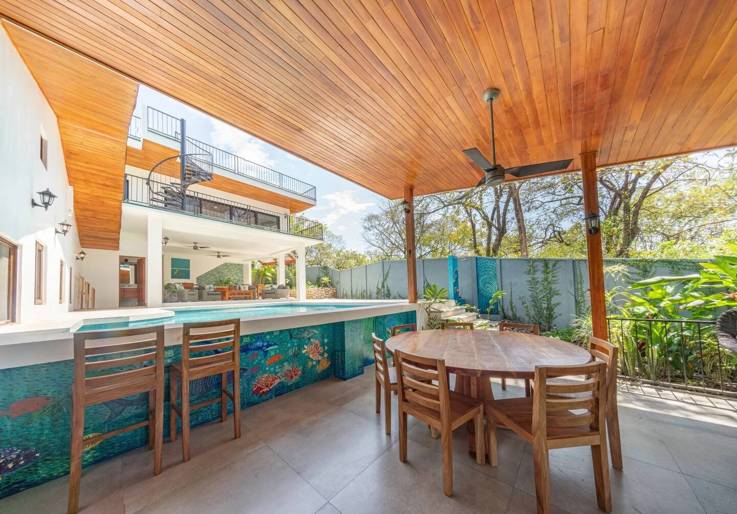 Covered patio with wooden ceiling, outdoor dining table with chairs, swimming pool with marine-themed mosaic, and backyard with trees and greenery.