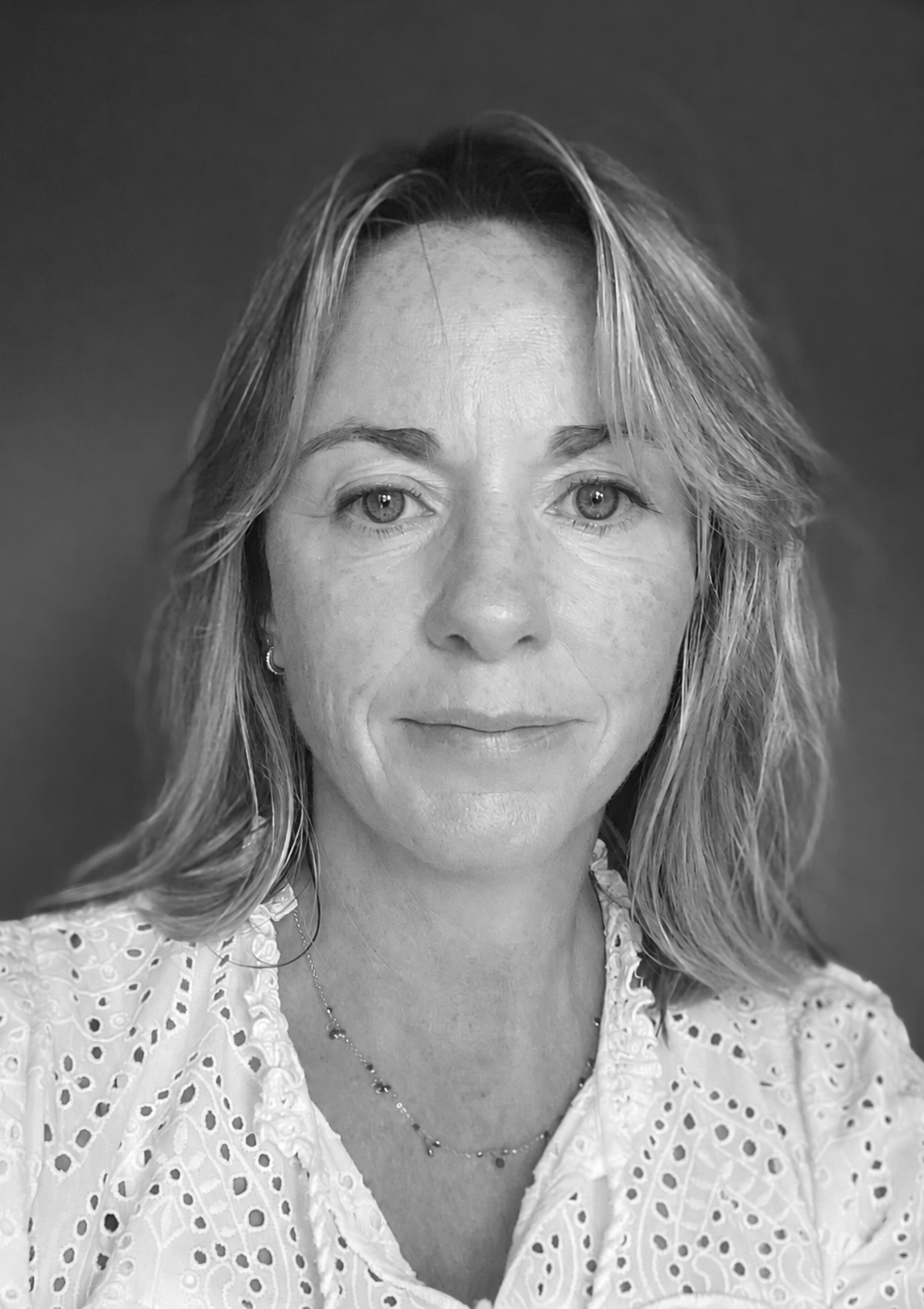 A black and white close-up portrait of a woman with shoulder-length wavy hair, wearing a white eyelet blouse and a delicate necklace, against a plain dark background.