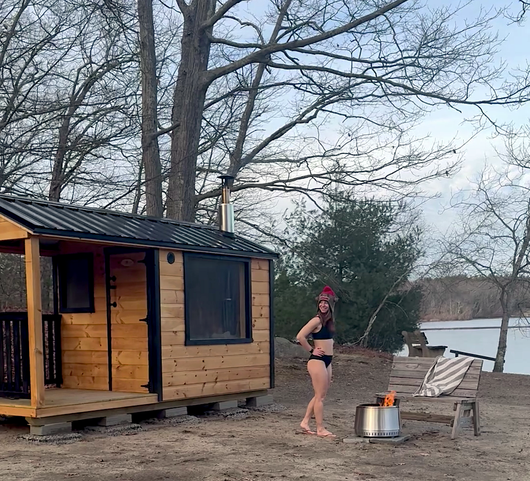 A woman in a black bikini and red knit hat standing outdoors near a small wooden cabin, with a fire pit and a lounge chair, by a lake with bare trees in the background.
