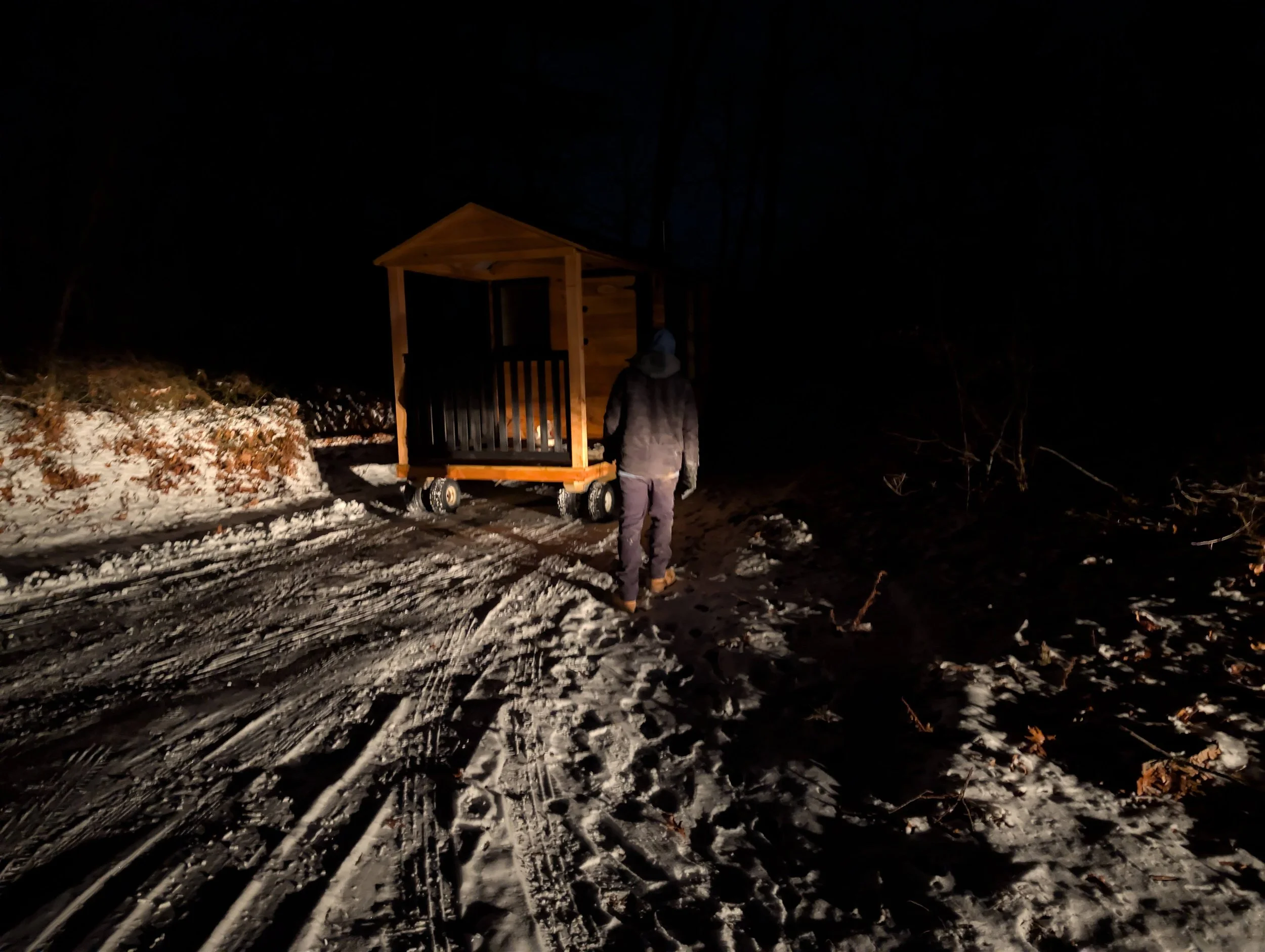 Person in winter clothing operating a small wooden mobile structure in a snowy, dark outdoor setting at night.