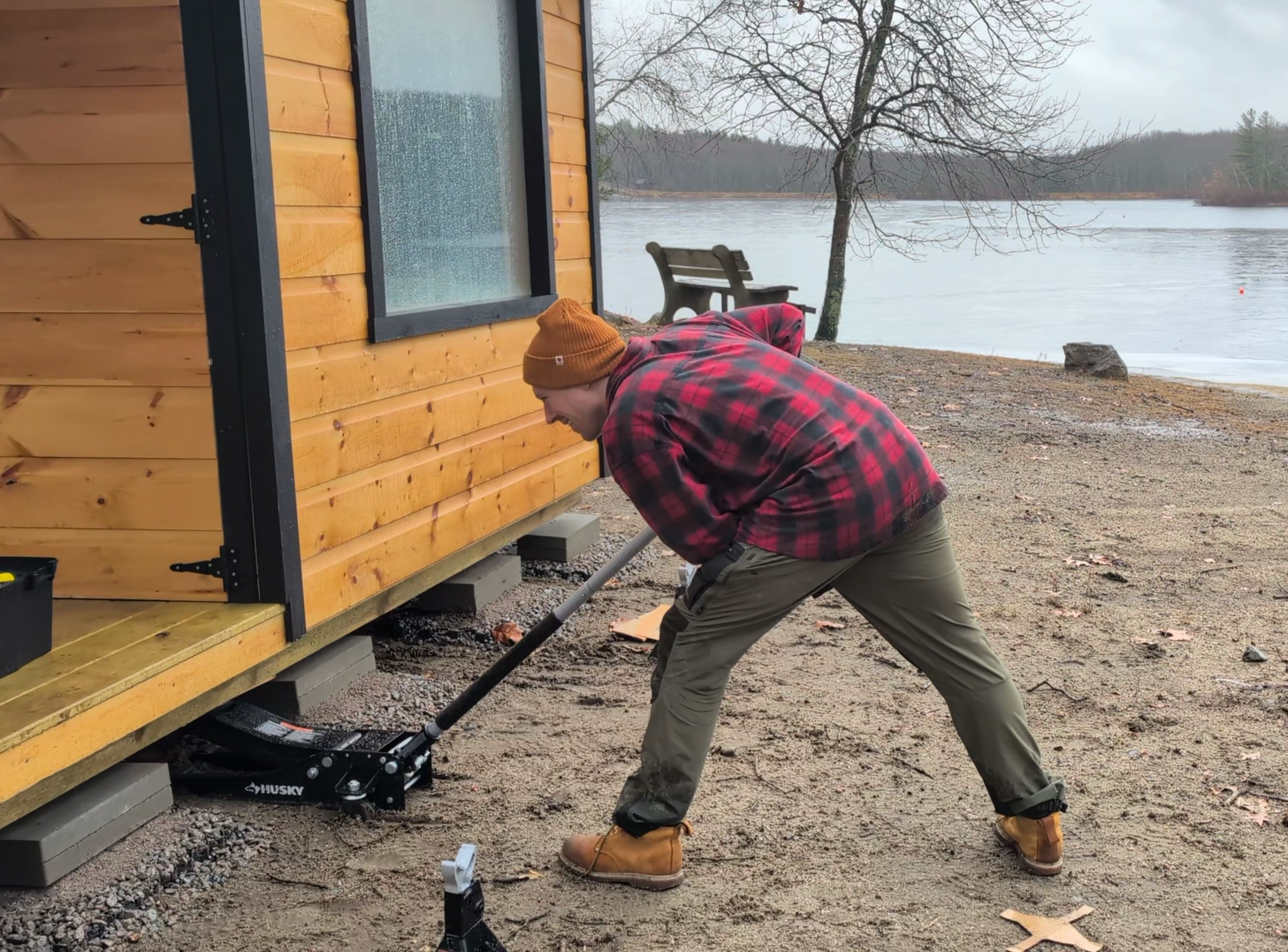 A person in a red and black plaid jacket and brown beanie using a hydraulic jack to lift the corner of a small wooden cabin on concrete blocks by a lakeside.