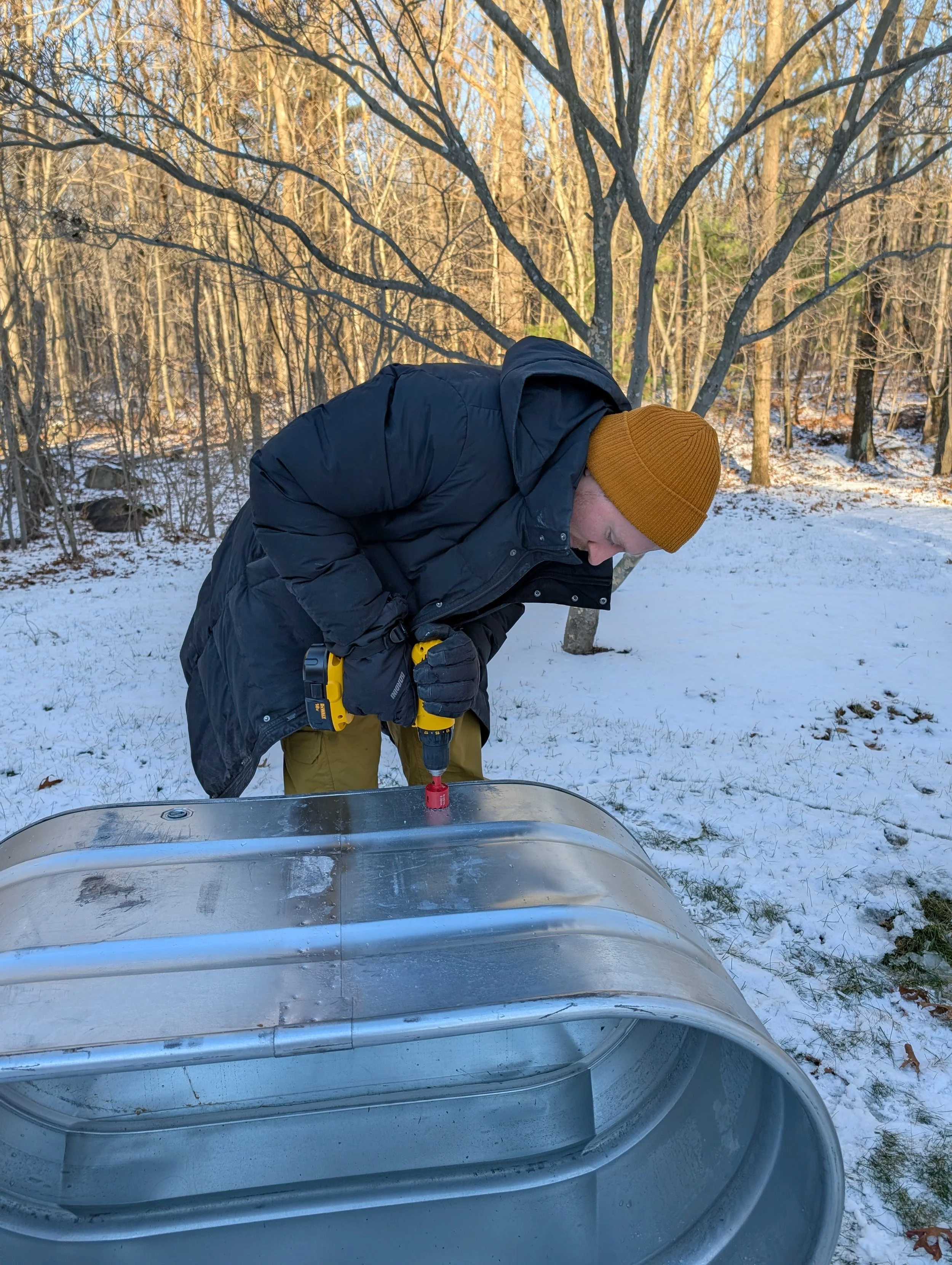 A person wearing a black jacket, mustard-colored beanie, and gloves working outdoors with power tools on a metal object, in a snowy, wooded area.