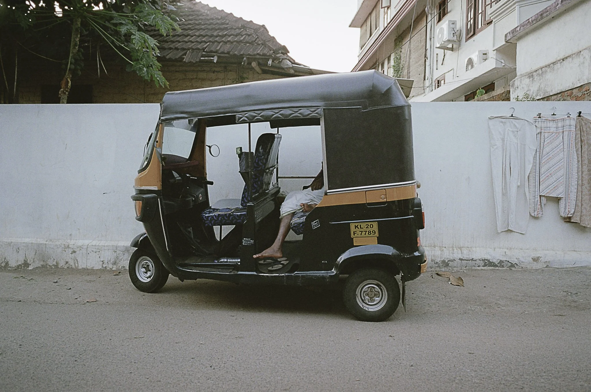 Tuk-tuk nero e arancione parcheggiato sulla strada, con una persona seduta all'interno con le gambe fuori e distesa, sullo sfondo un muro bianco con alcuni vestiti appesi e case in fronte