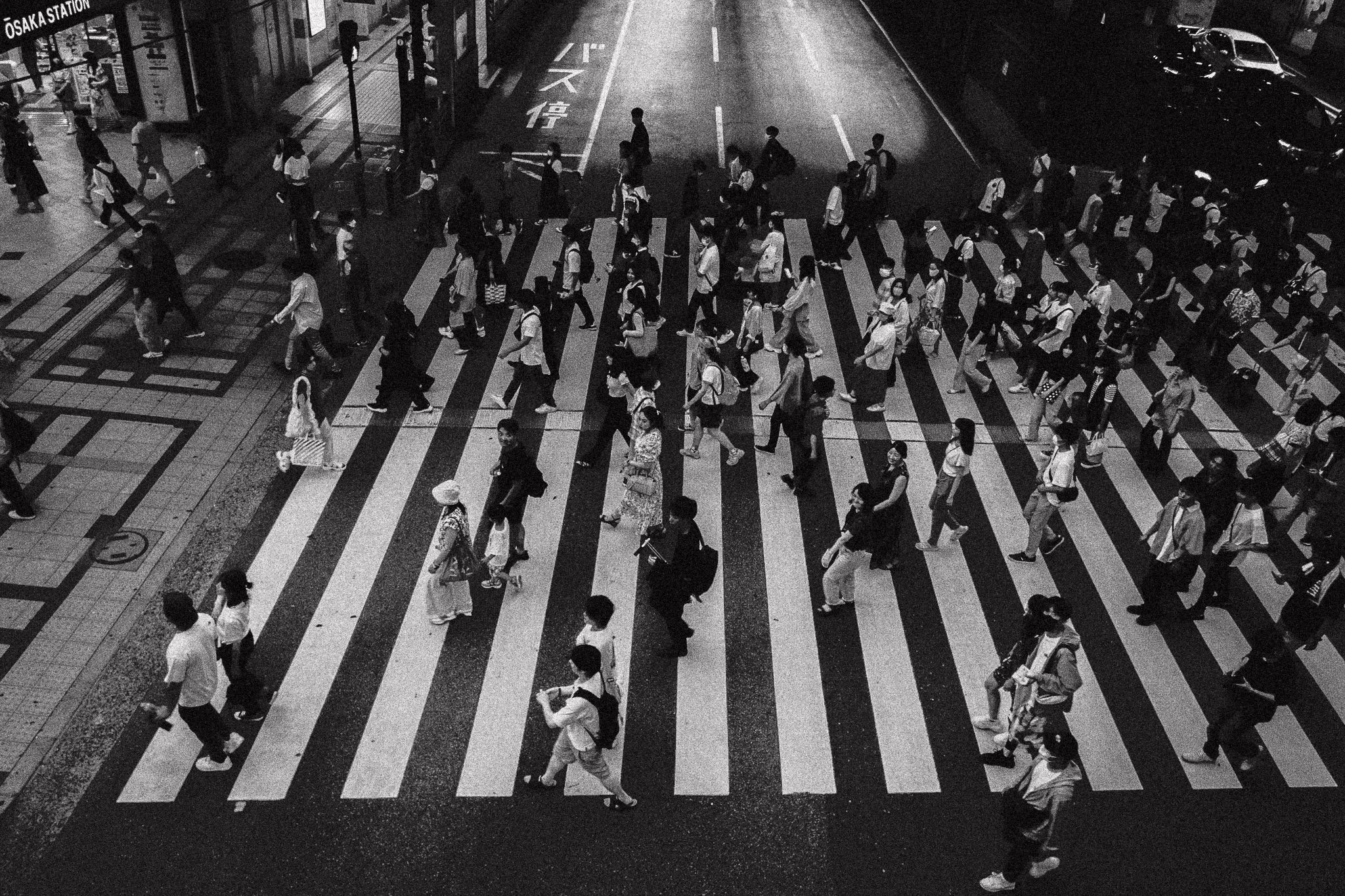 Persone che attraversano un pedonale affollato in città durante la notte, con luci e negozi visibili ai lati della strada.