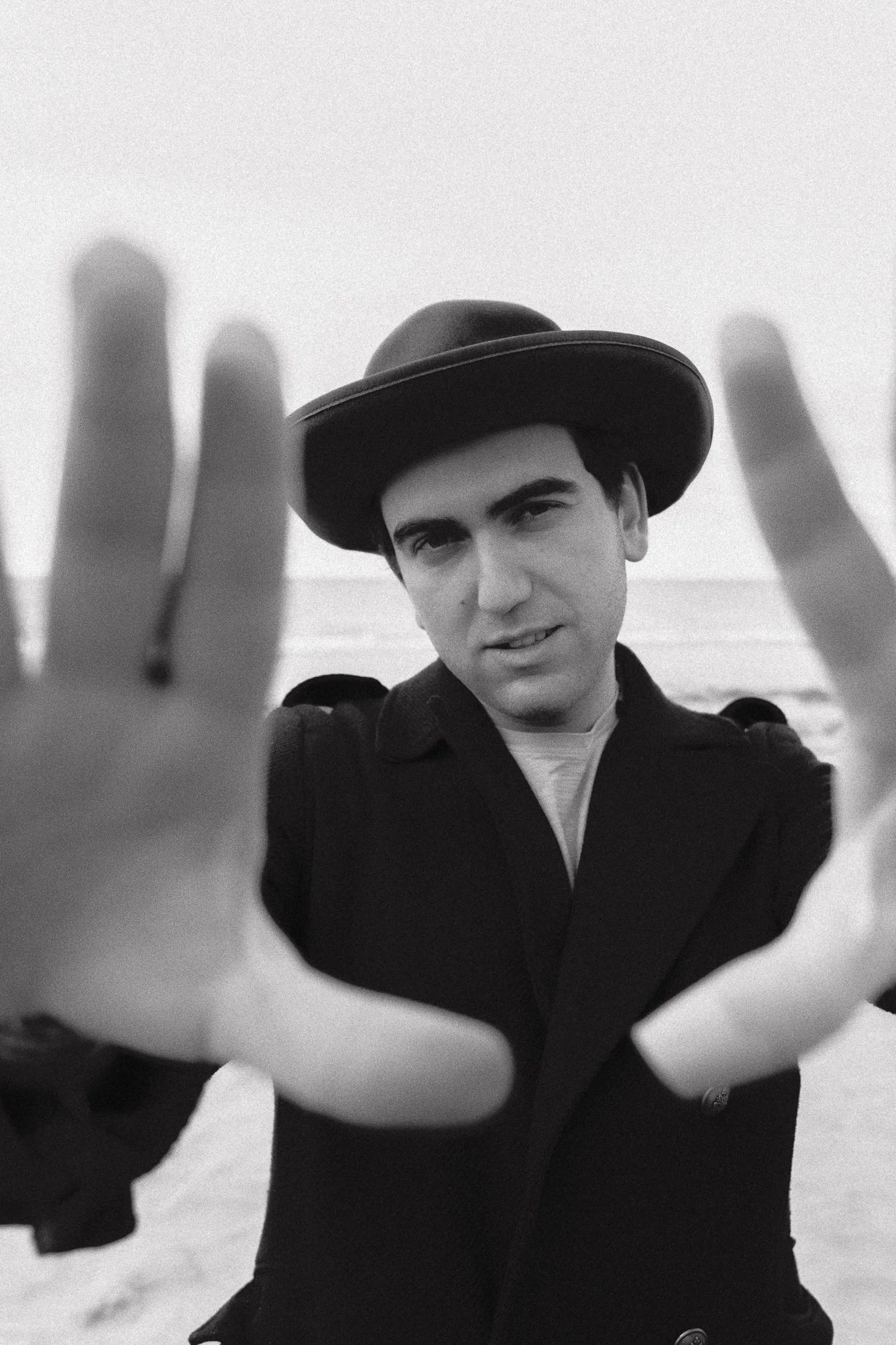 Un uomo che indossa un cappello in stile cowboy e una giacca scura, si avvicina alla fotocamera con le mani aperte, sullo sfondo sembra esserci il mare