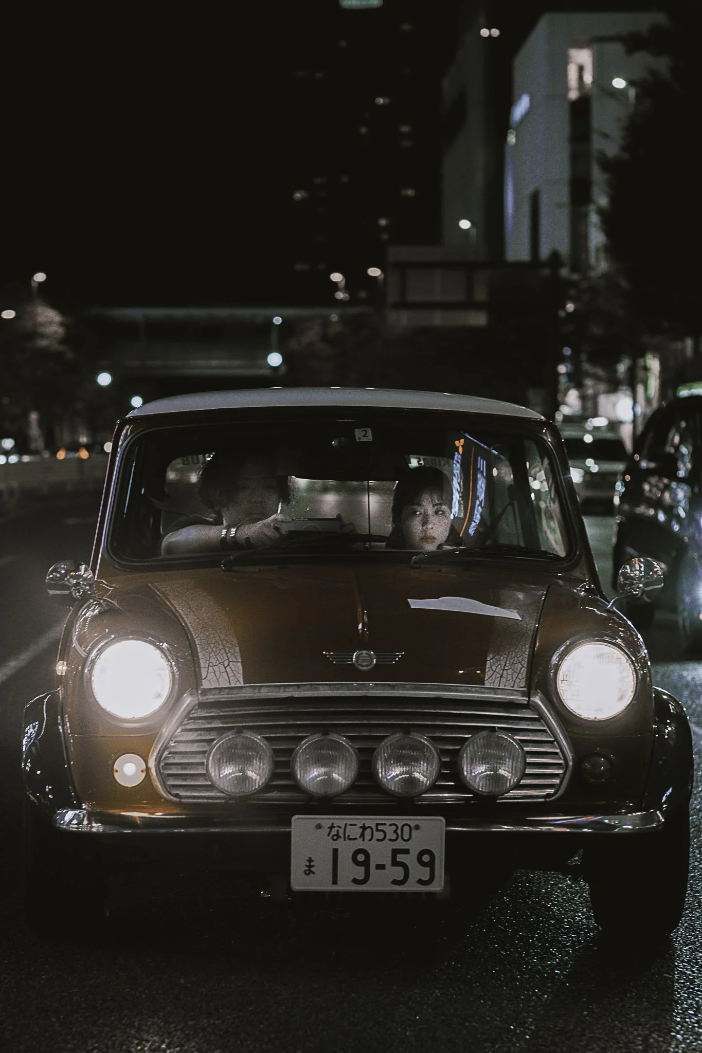 Una macchina d'epoca rossa con quattro fari supplementari, in una strada illuminata di notte, con due persone visibili all'interno.