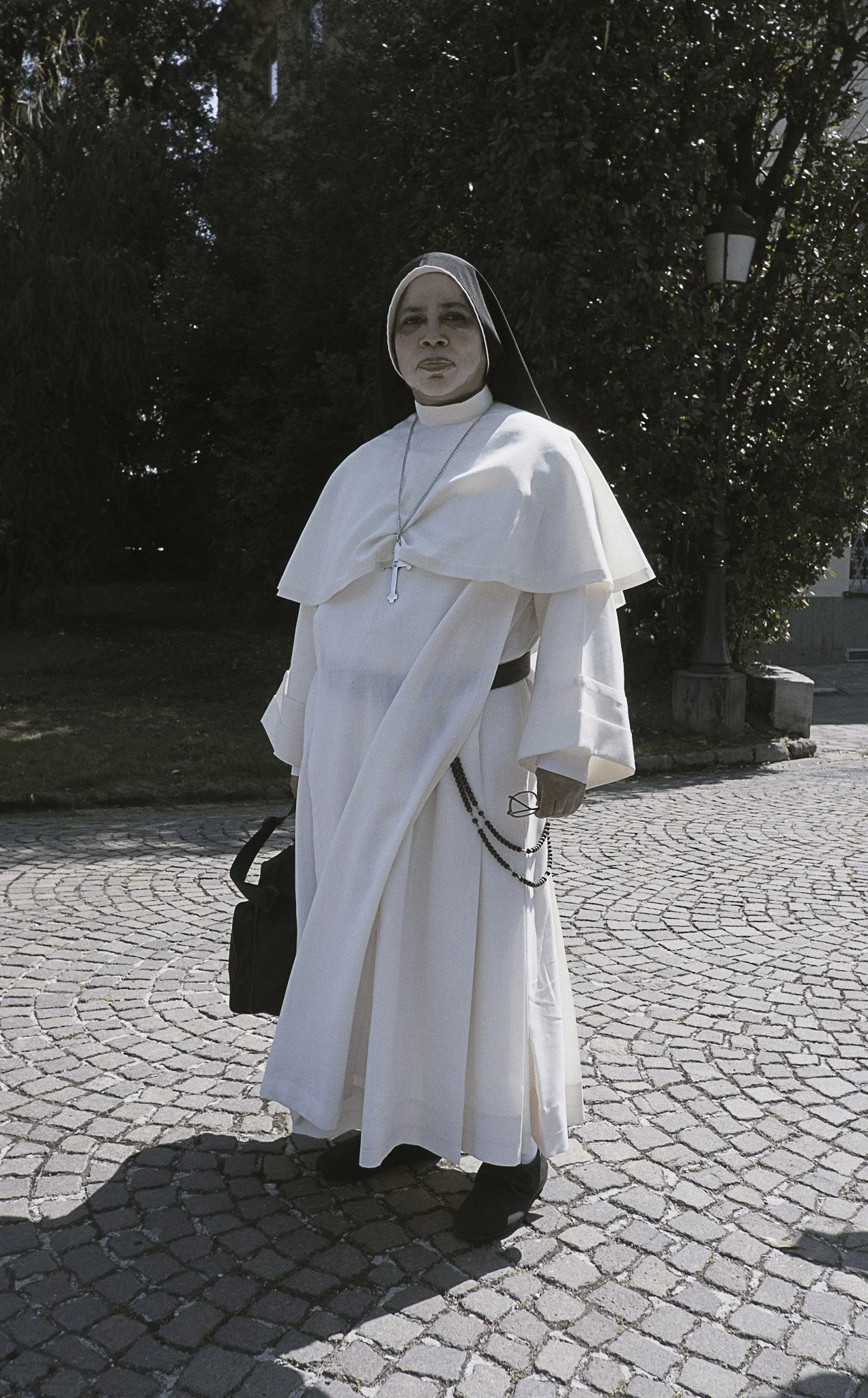 Persona con abiti religiosi, indossa un abito bianco con una croce e un rosario, situata in una piazza con pavimentazione in ciottoli e alberi sullo sfondo.