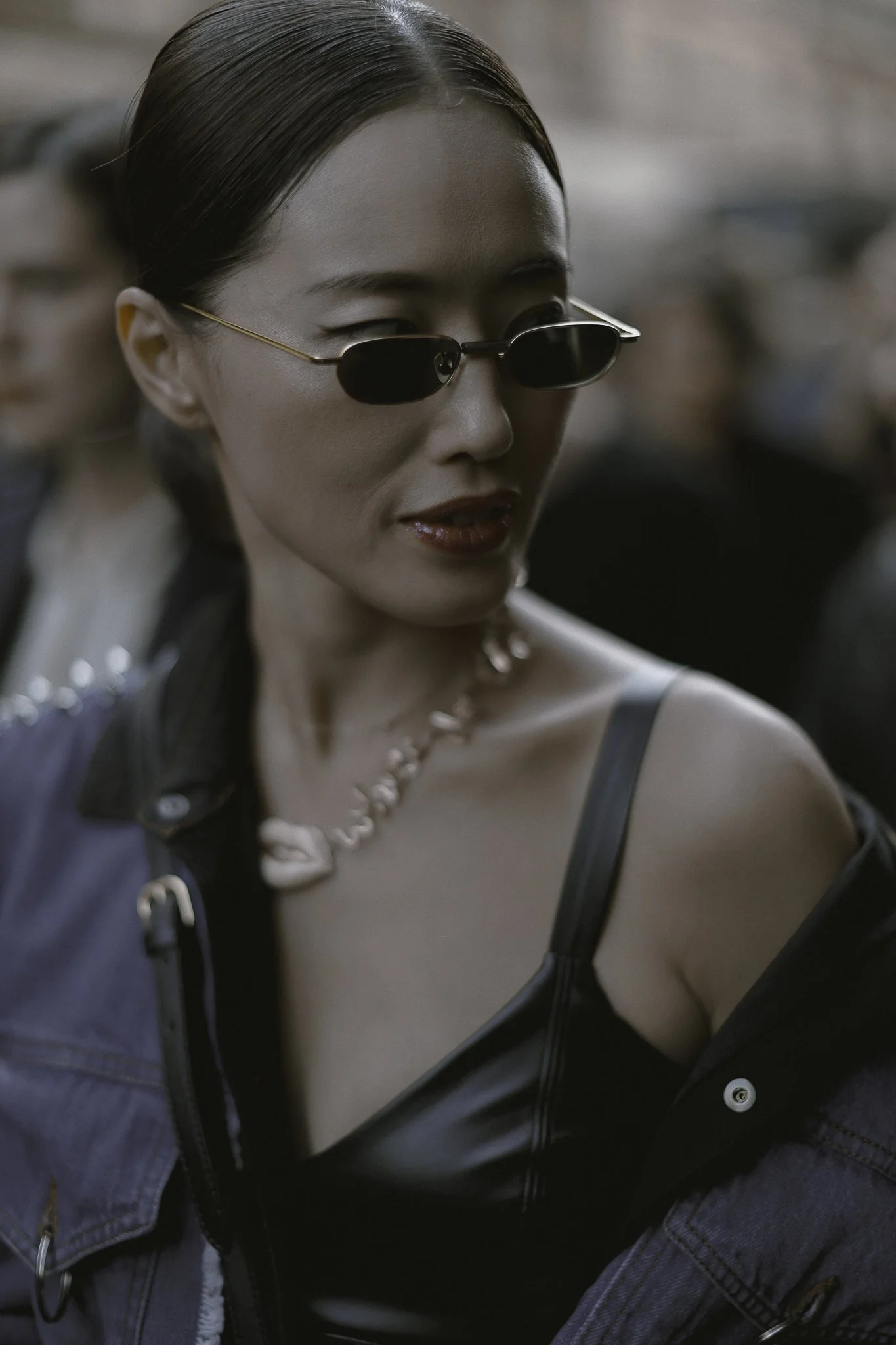 Donna con occhiali da sole neri, capelli raccolti, abbigliamento alla moda e accessori, in un ambiente urbano.