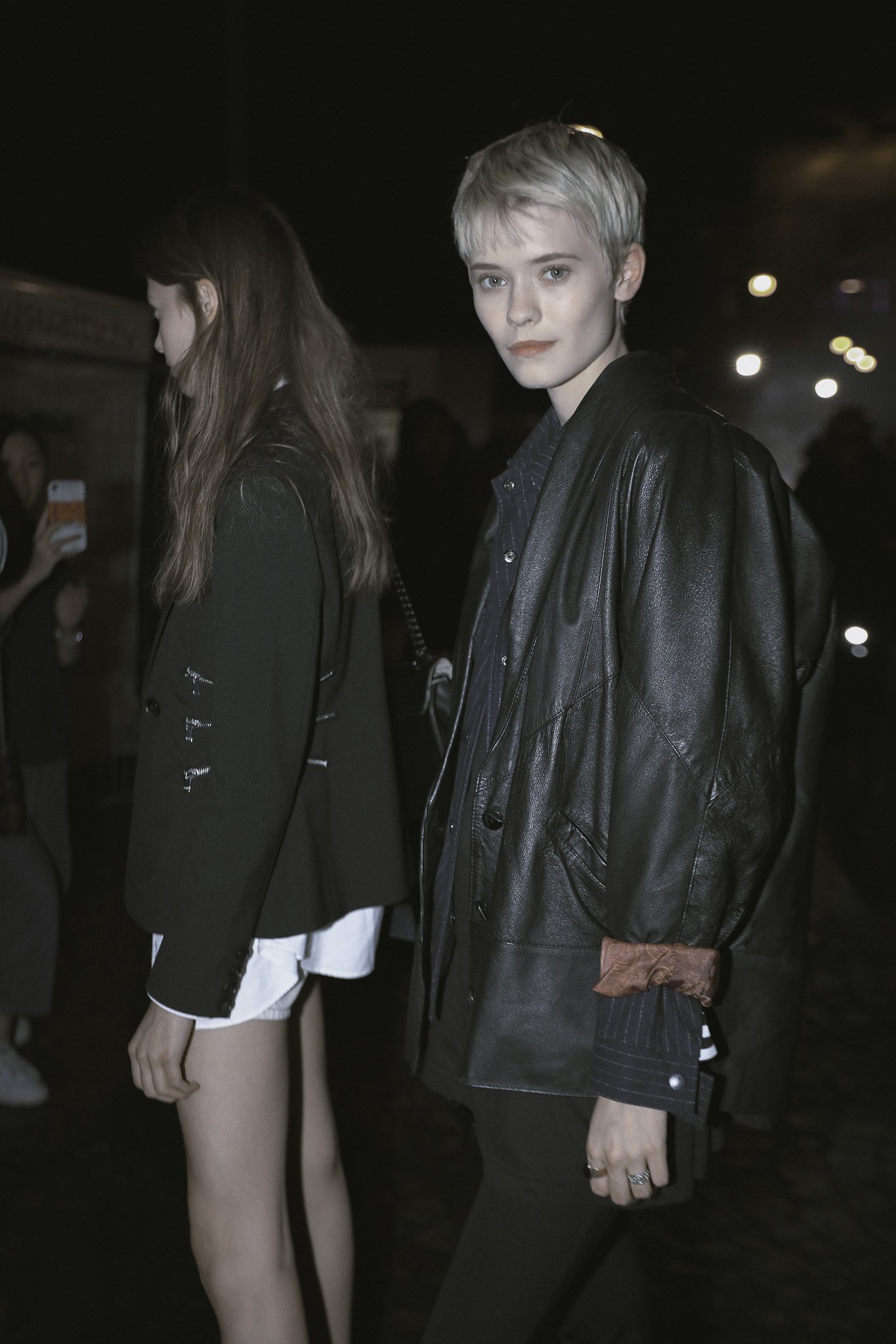 Una giovane donna con capelli corti e biondi chiusi in una giacca di pelle nera, con una camicia a righe e un anello, guarda verso la telecamera in un ambiente con luci soffuse e altre persone sullo sfondo.