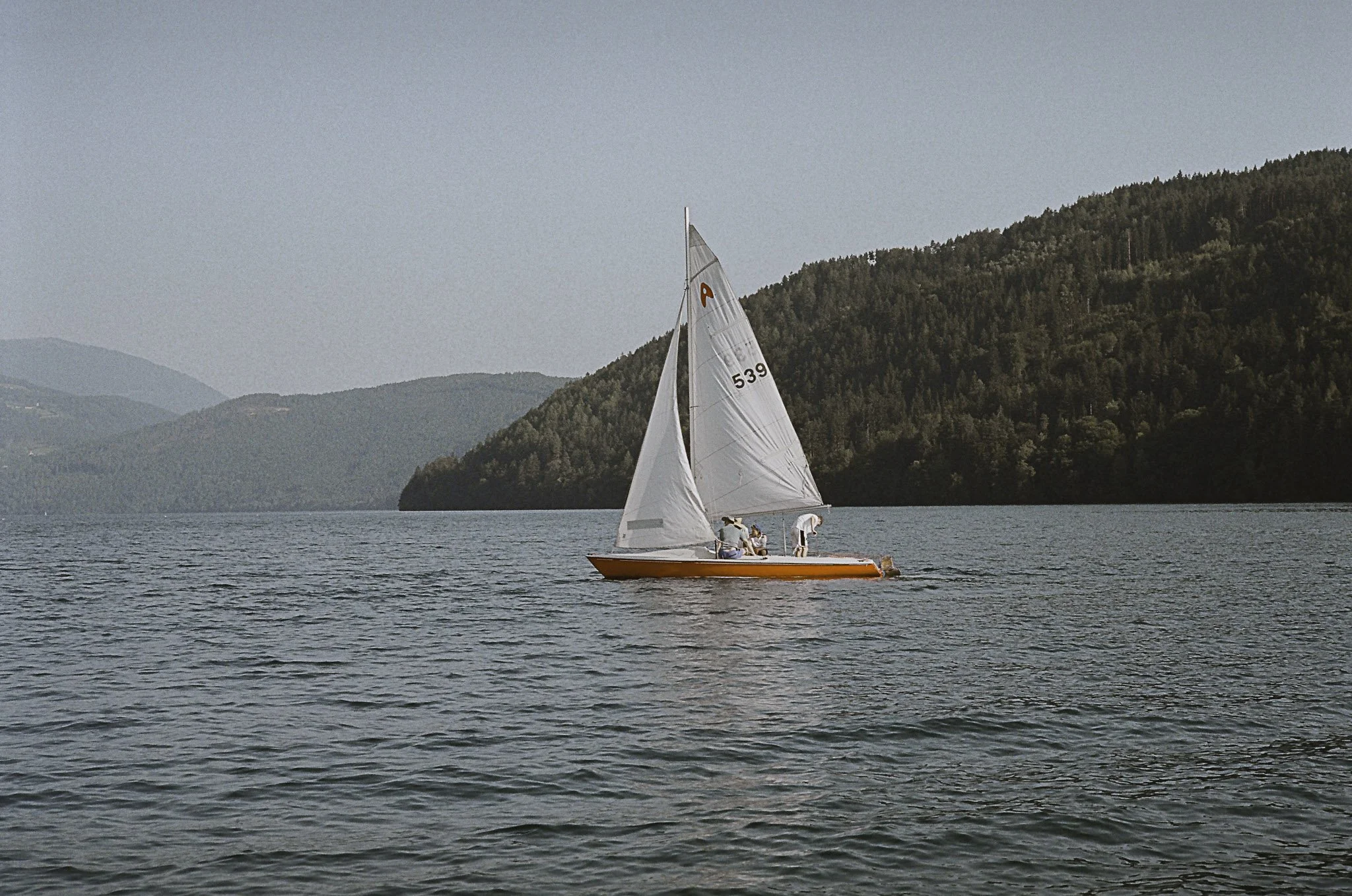 Una barca a vela con persone a bordo in acque calme, con colline boschive sullo sfondo