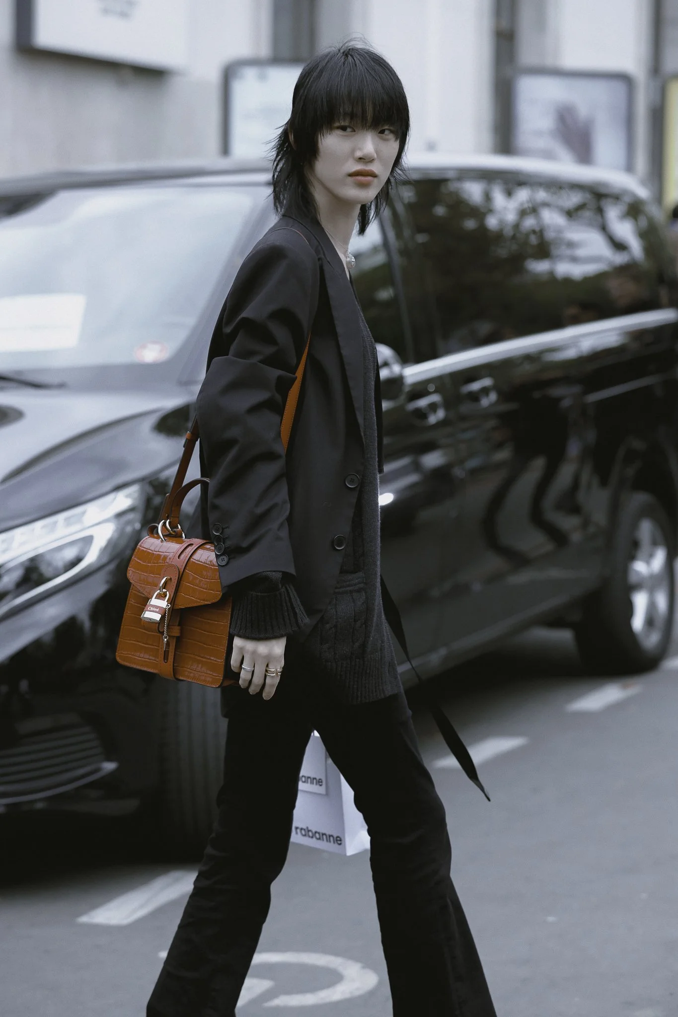 Una donna con capelli neri corti, vestita con un abbigliamento nero, attraversa la strada con una borsa rossa a tracolla, accanto a un'auto nera parcheggiata in città.