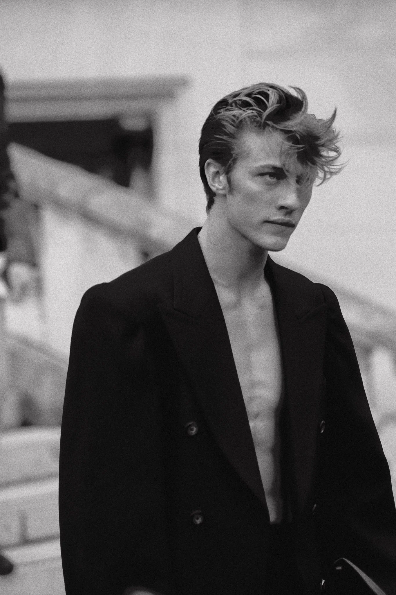 Ragazzo con capelli spettinati e senza maglietta sotto un cappotto nero, in bianco e nero.