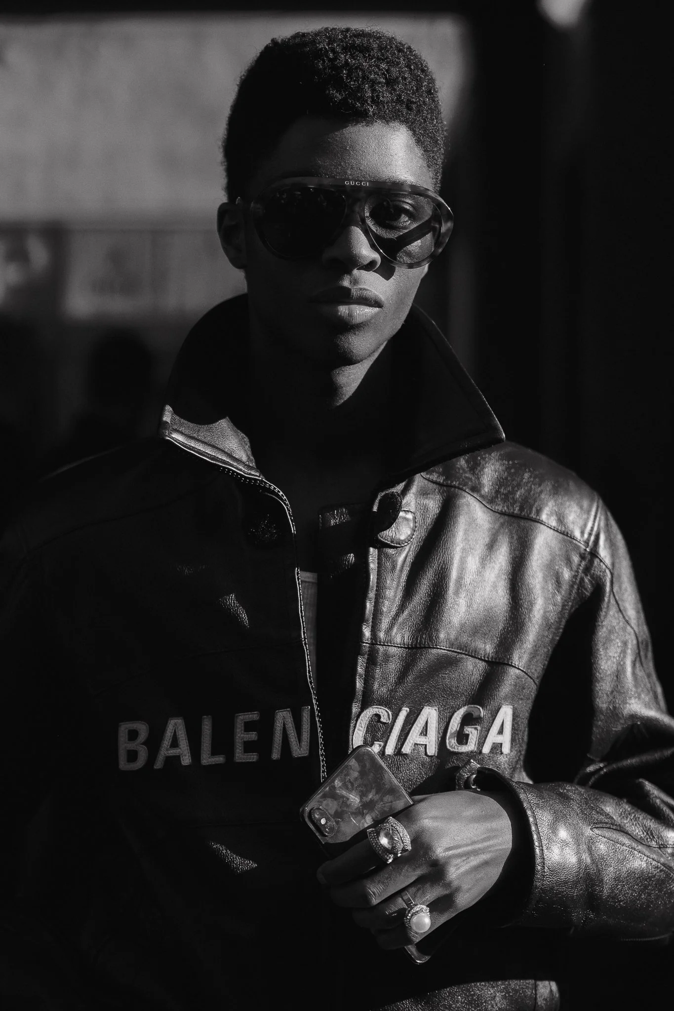 Ritratto in bianco e nero di una donna con pelle scura, occhiali da sole Gucci, cappotto in pelle con la scritta Balenciaga, che tiene uno smartphone con una mano e indossa diversi anelli.