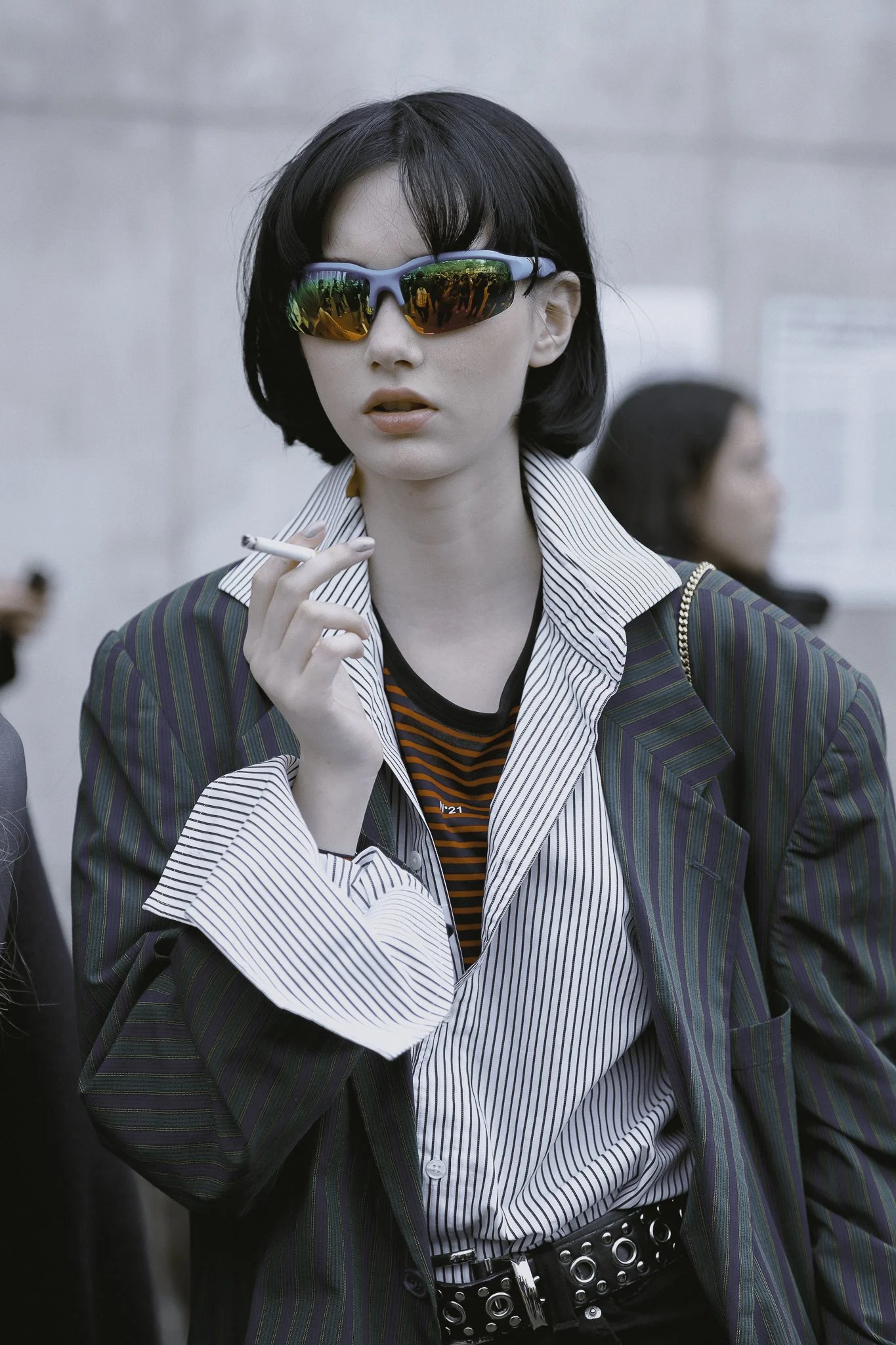 Donna con capelli corti e occhiali da sole riflettenti, indossa una giacca a righe e fuma una sigaretta