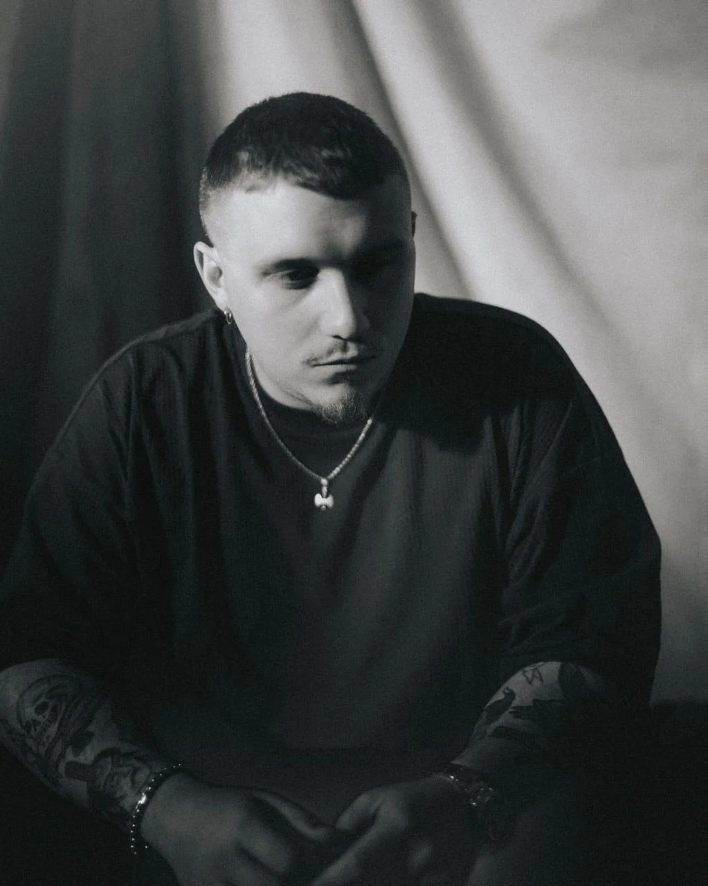 Ritratto in bianco e nero di un giovane uomo con capelli corti, tatuaggi visibili sulle braccia, orecchino e collana, in un ambiente con sfondo sfocato e illuminazione soffusa.