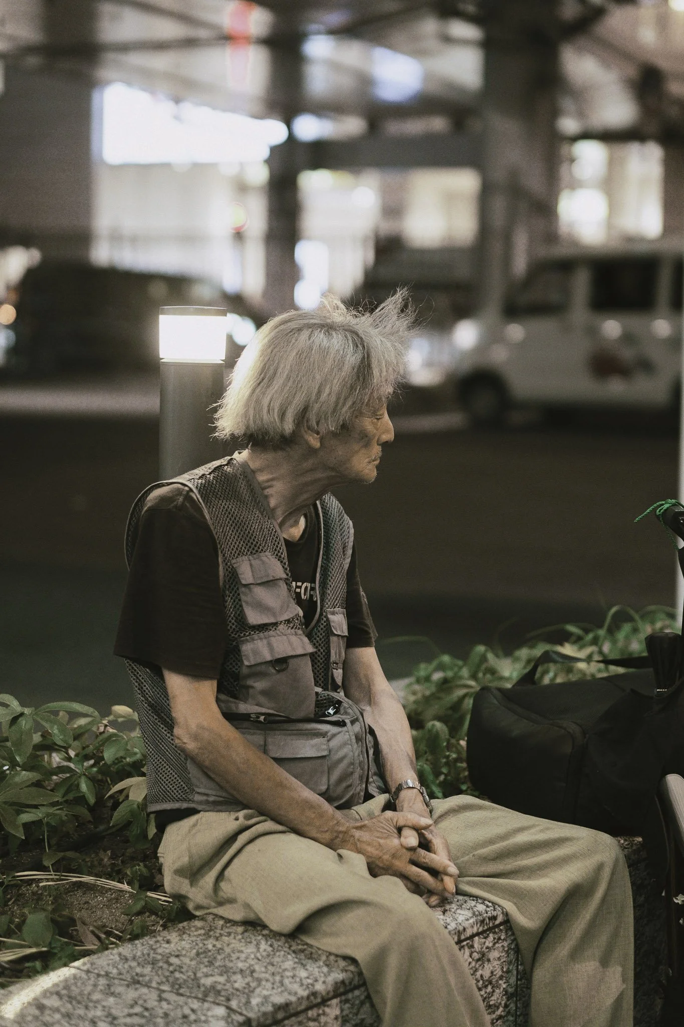 Una donna anziana seduta su una panchina di sera o notte, in un'area pubblica con luci soffuse e veicoli in movimento sullo sfondo.