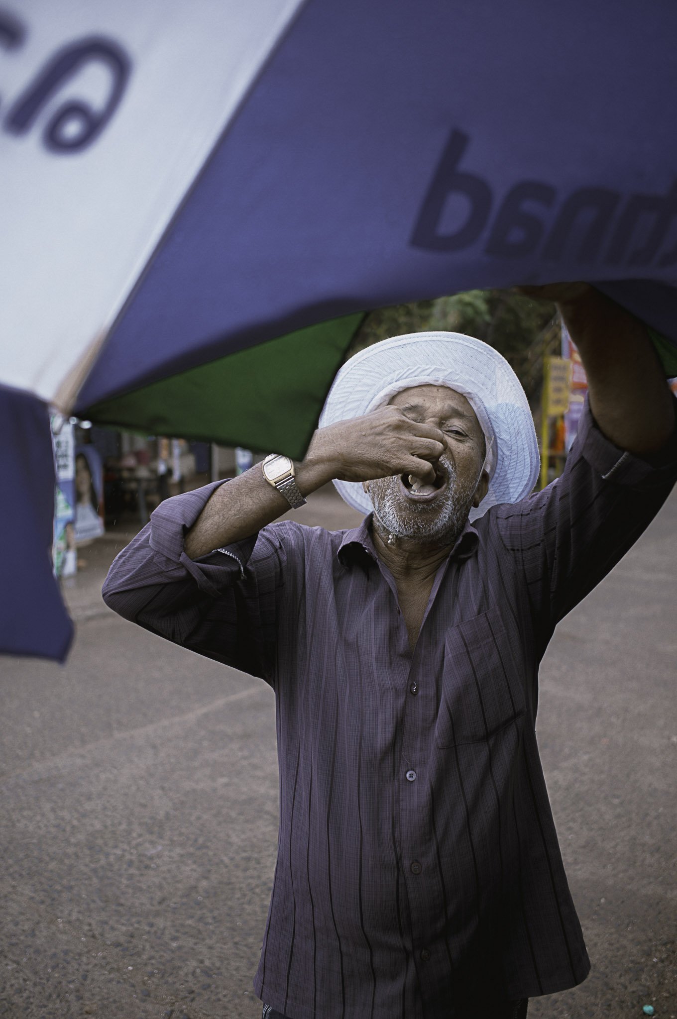 Uomo anziano con cappello bianco si pulisce il naso con il dorso della mano sotto un ombrello di colore blu e verde, sullo sfondo una strada con alcune bancarelle.