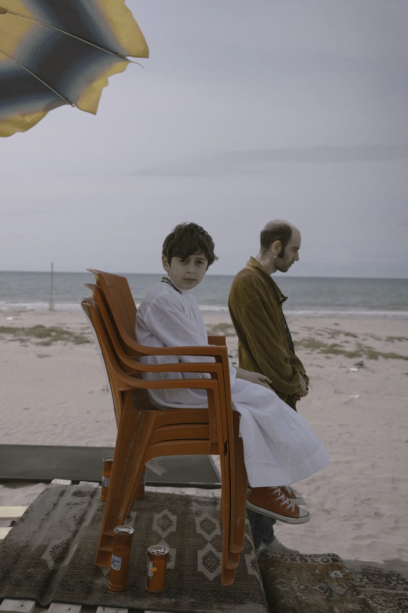 Due persone, un ragazzo e un uomo, seduti su sedie di plastica arancioni sulla spiaggia, con il mare sullo sfondo. Ci sono birre sul pavimento davanti a loro.