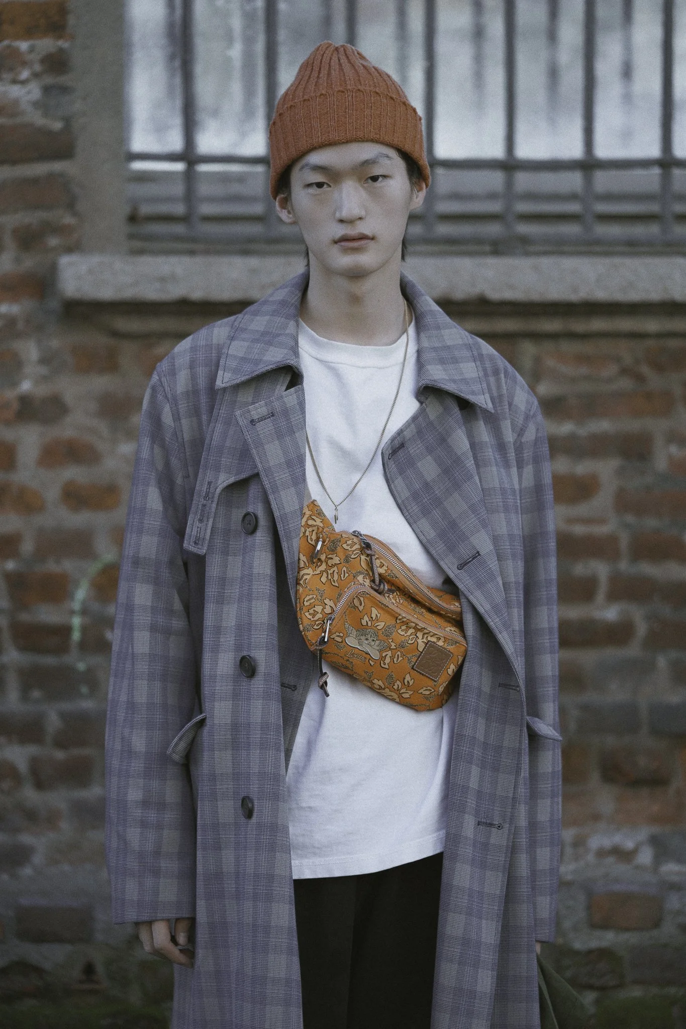 Ragazzo con cappello di lana arancione, giacca lunga a quadri grigia, maglietta bianca, portafoglio da cintura arancione con disegni floreali, in piedi davanti a un muro di mattoni.