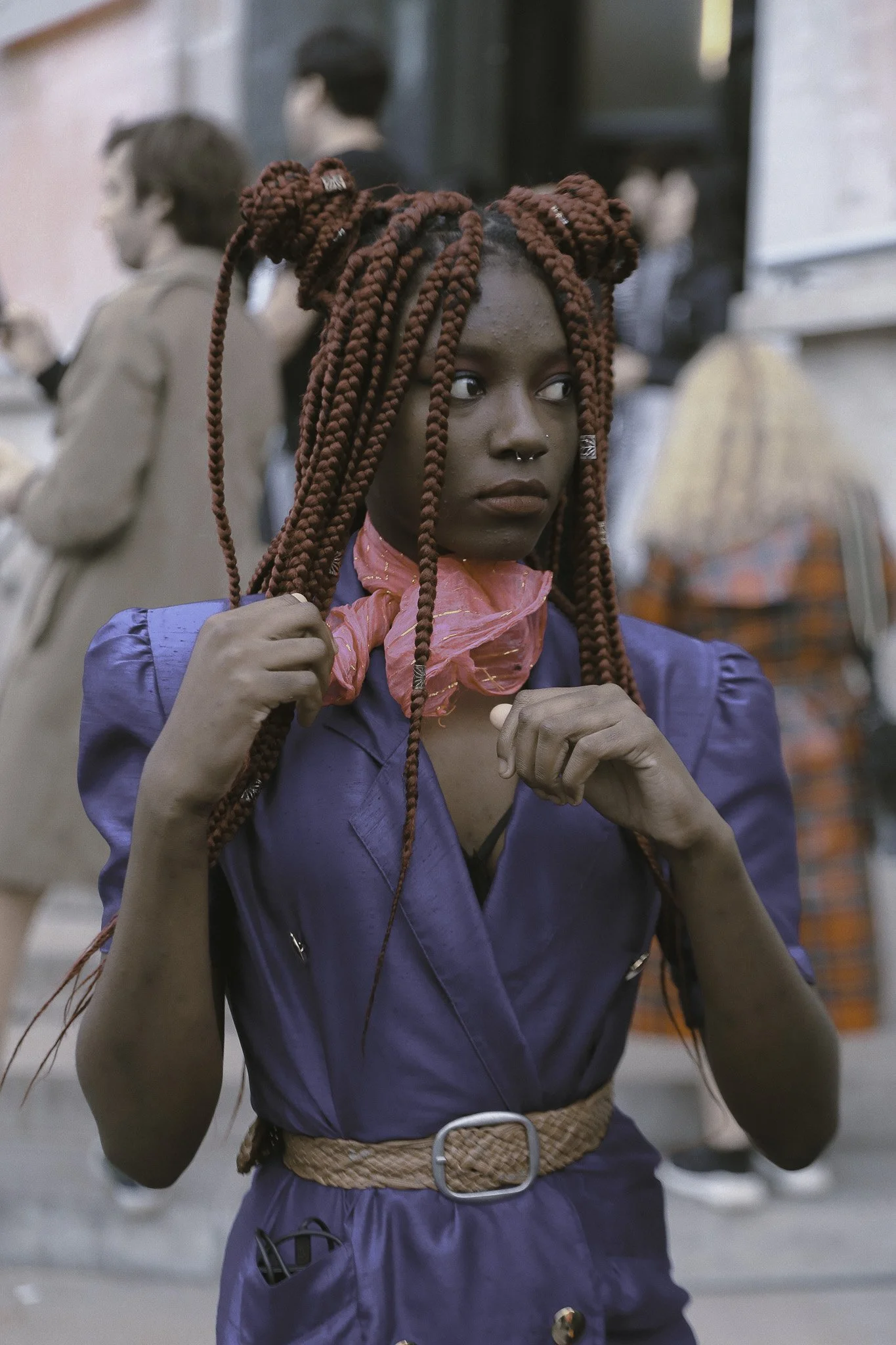 Donna con trecce rosse e pelle scura indossa un vestito blu, una sciarpa rosa e si trova in una strada affollata.
