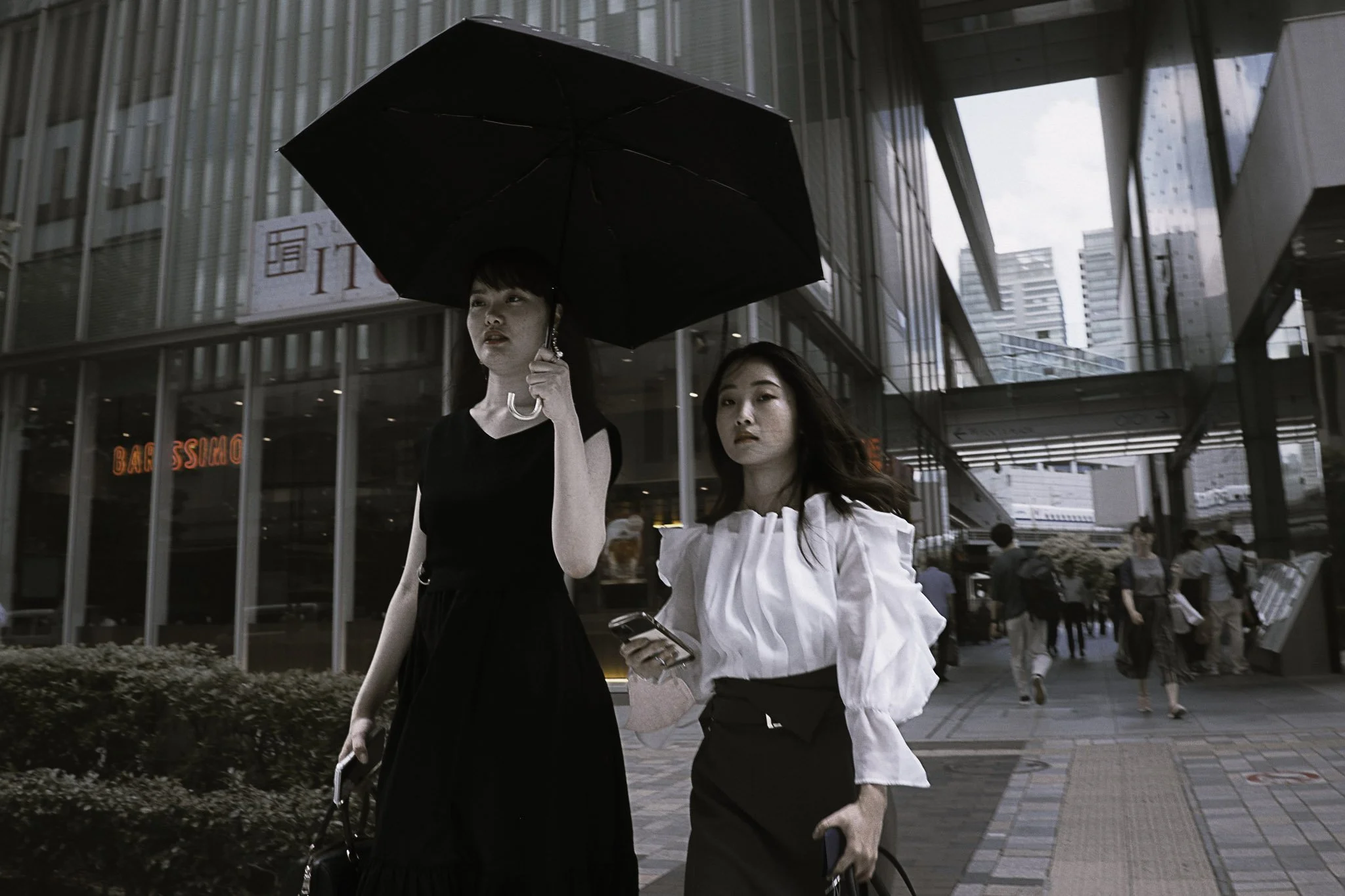 Due donne camminano per strada sotto un ombrello nero, una con un vestito nero e l'altra con una camicia bianca con maniche a sbuffo e una gonna scura, in un'area urbana moderna con edifici di vetro.