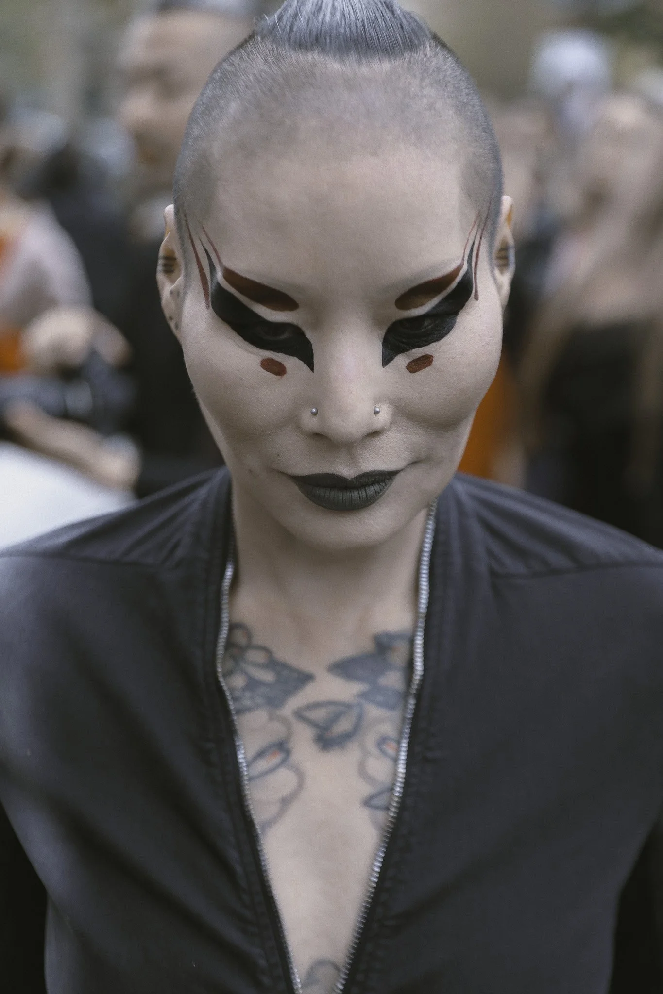 Persona con trucco e tatuaggi, che indossa una giacca nera, in un ritratto in primo piano.