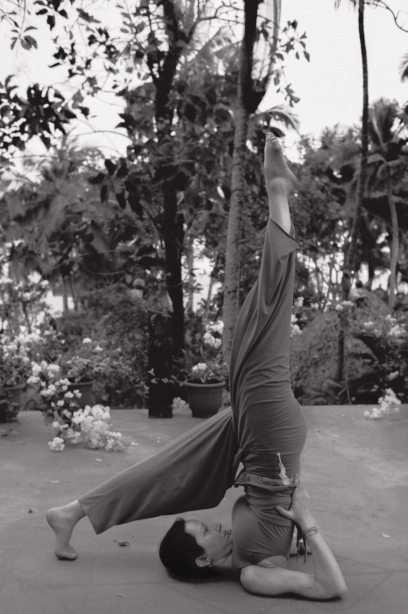 Una donna fa yoga all'aperto, in posizione di equilibrio sulla testa con le gambe alzate in verticale, in un giardino con piante e fiori.