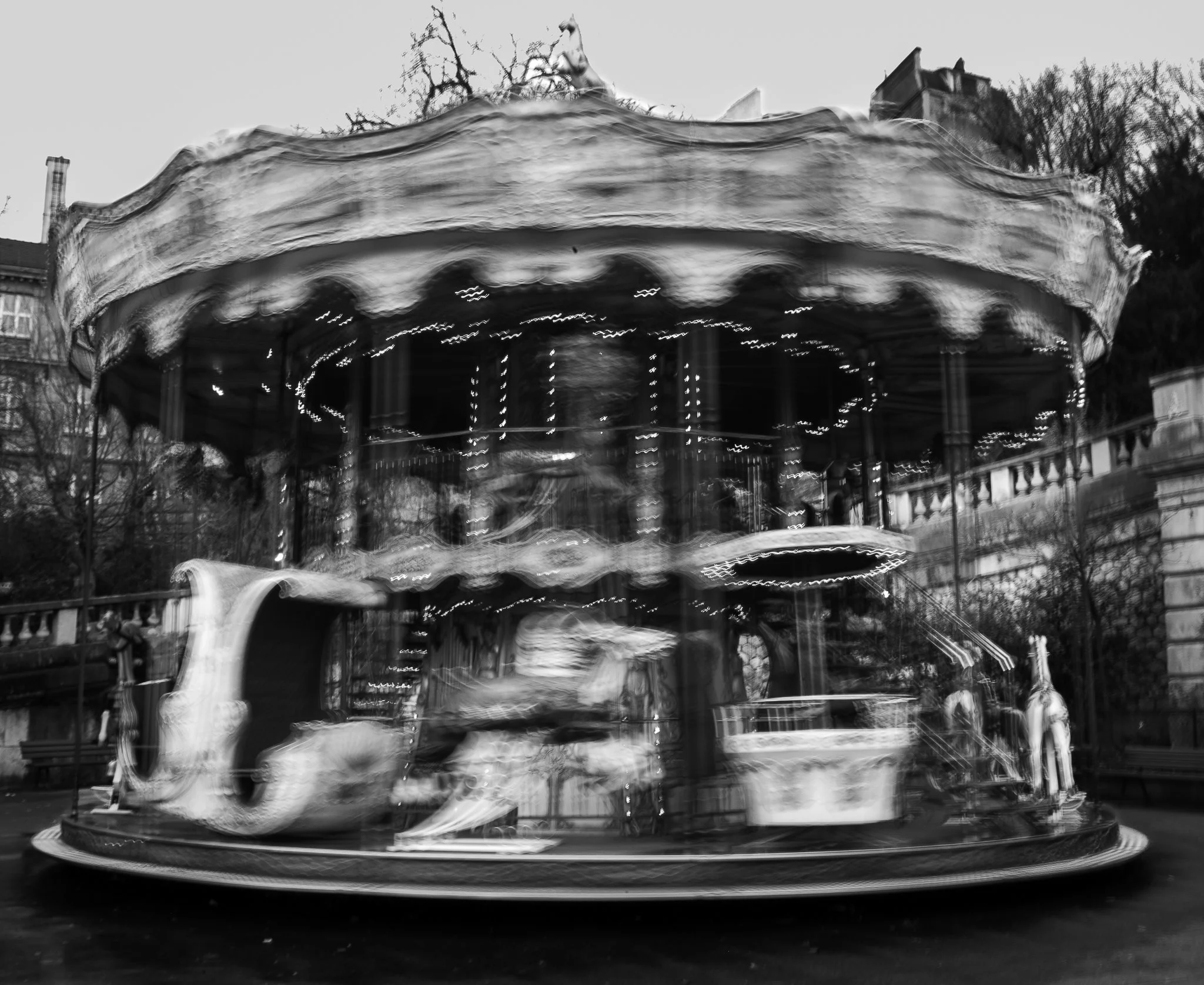 Giostra a due piani con luci decorative, in movimento, in un parco urbano. Sfondo di alberi e edifici, immagine in bianco e nero.