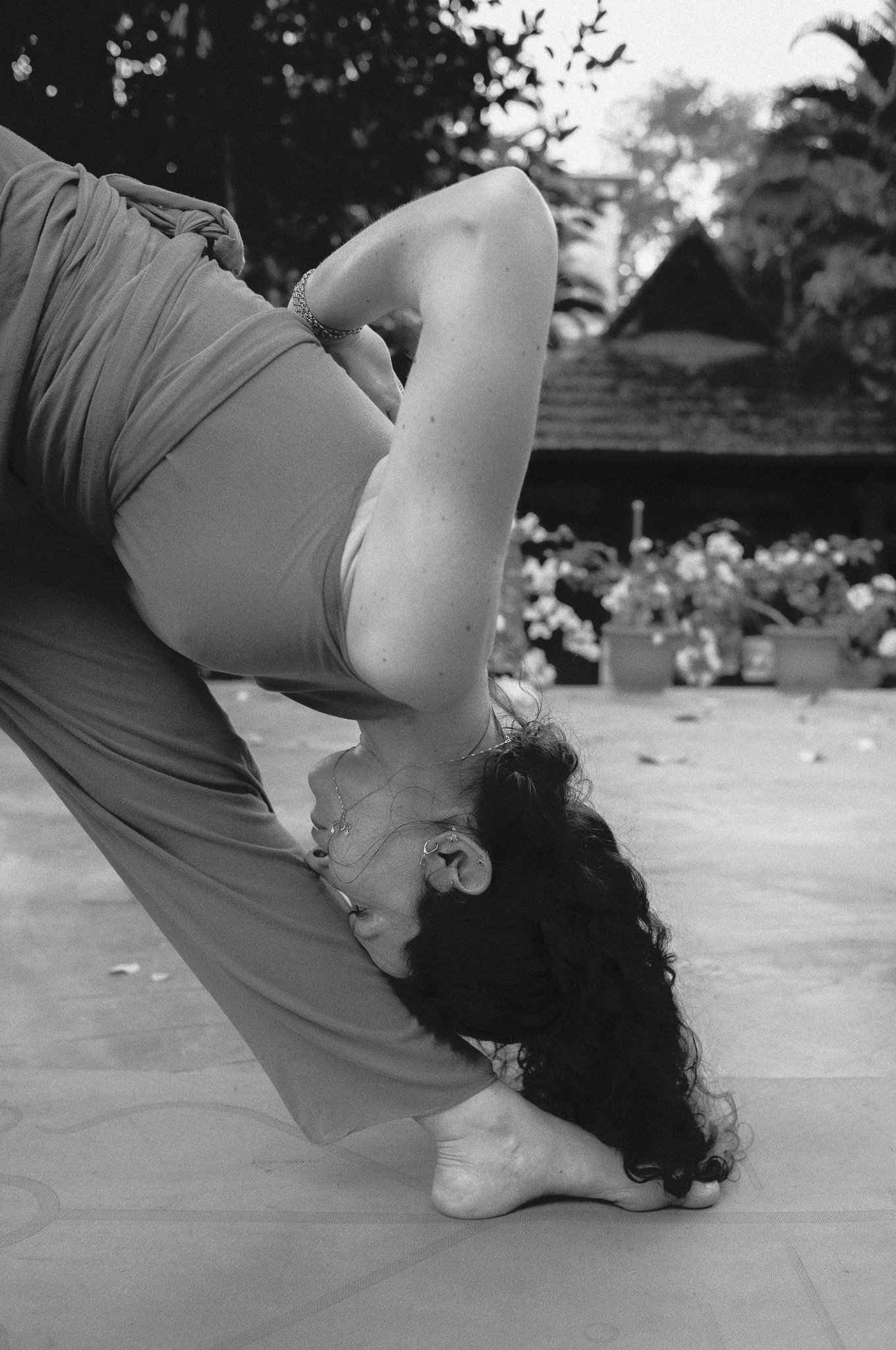 Una persona sta facendo una posizione yoga flessibile, con i capelli ricci neri, all'aperto, con fiori e piante sullo sfondo.