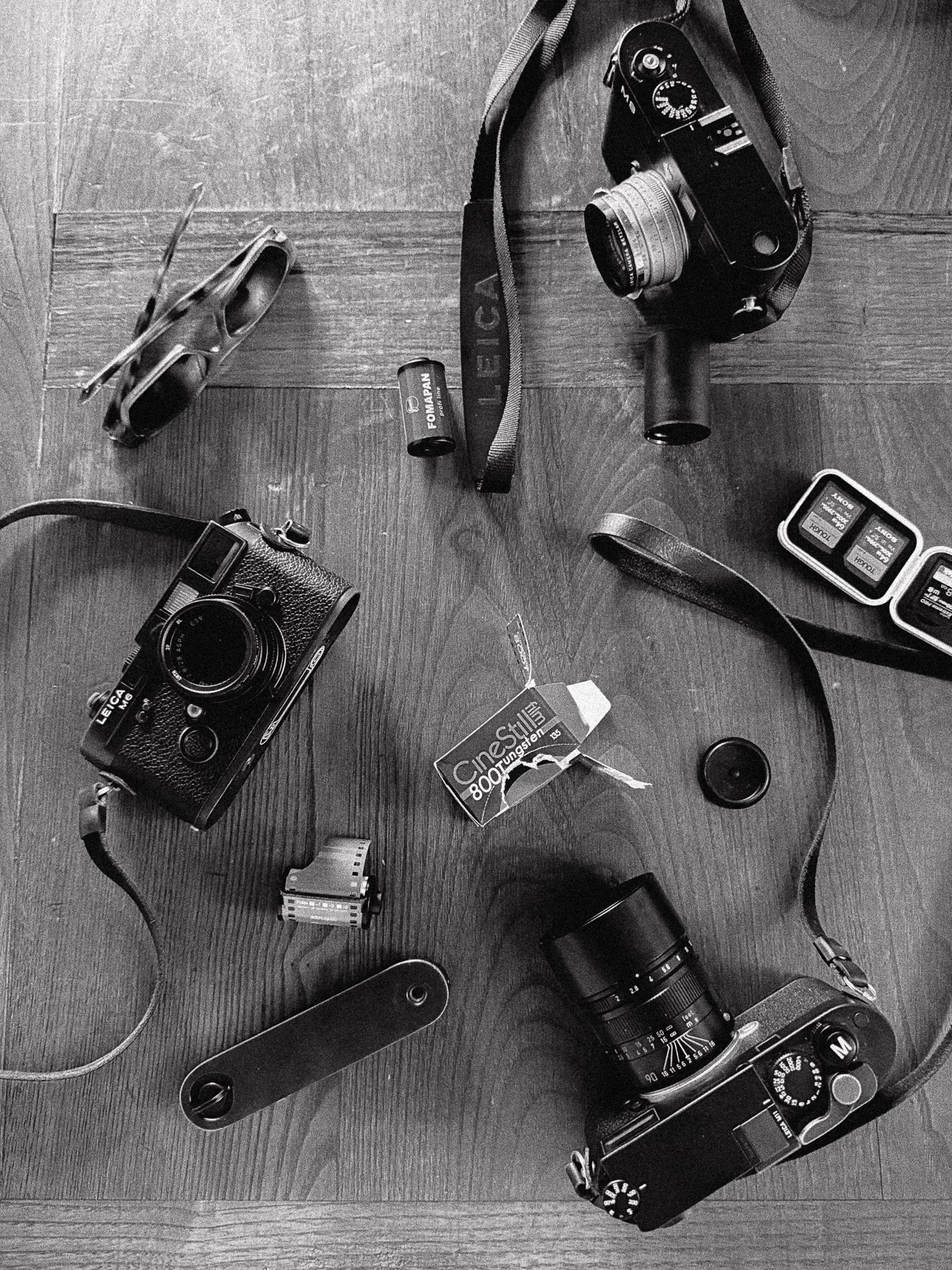 Vari attrezzature fotografiche disposte su un pavimento di legno: due fotocamere, obiettivi, batterie, schede di memoria, cacciaviti, e filtri fotografici, in un'immagine in bianco e nero.