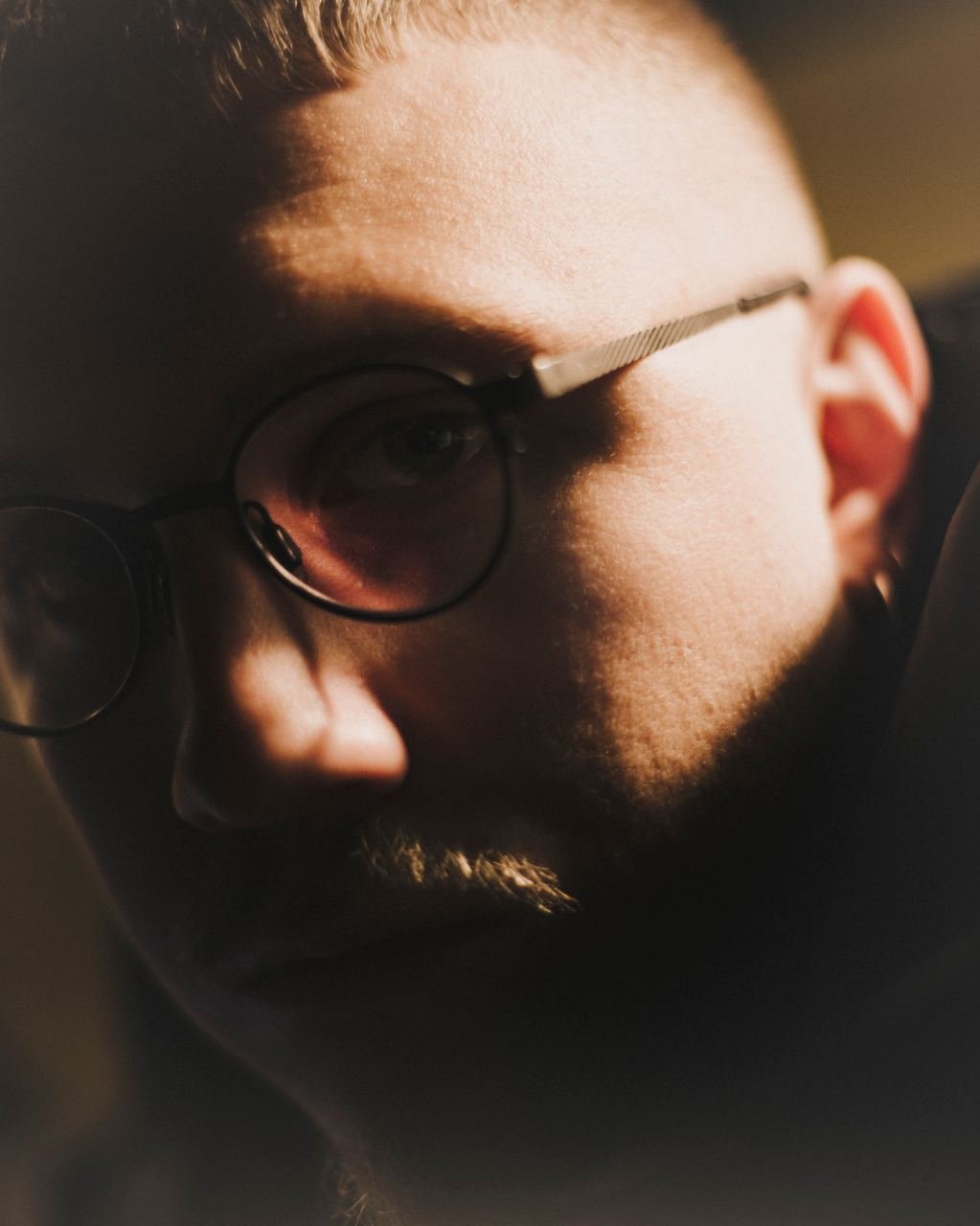 Primo piano di un uomo con occhiali tondi, barba e capelli corti, che utilizza un dispositivo elettronico, con illuminazione calda e sfondo sfocato.