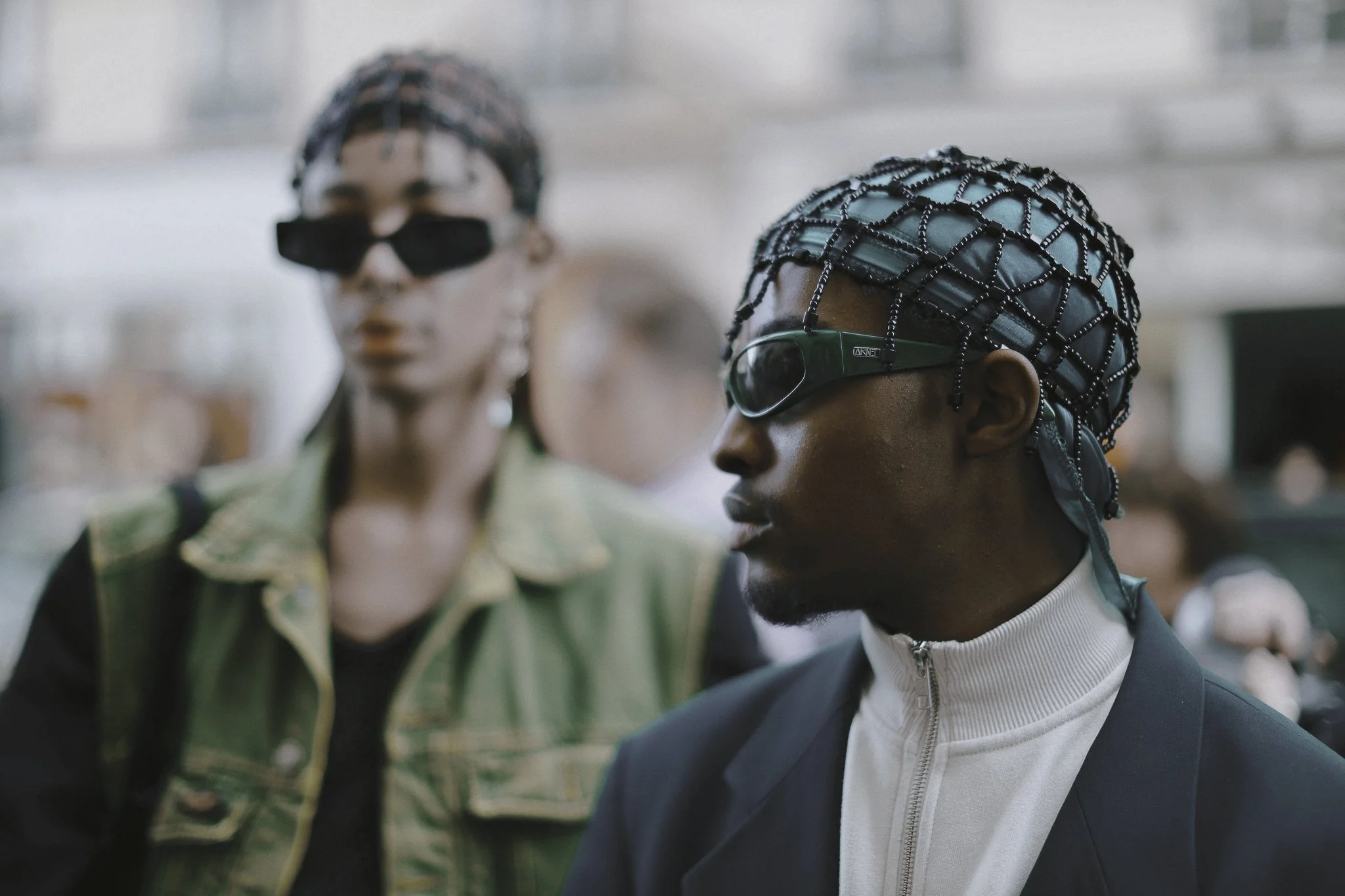 Due persone con capelli con intricati accessori e occhiali scuri, una ragazza in primo piano con a fianco un uomo, entrambi in un ambiente urbano.