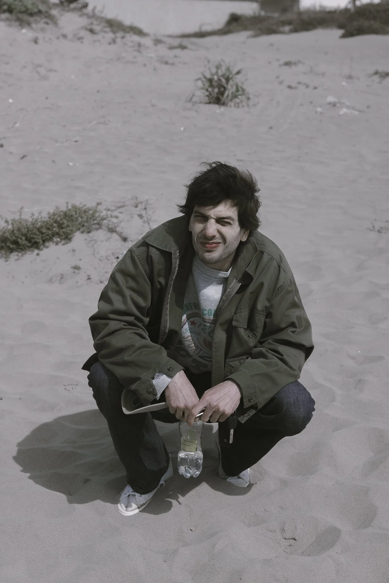Uomo con giacca verde in ginocchio sulla sabbia, che tiene una bottiglia di plastica con acqua, in spiaggia desertica.