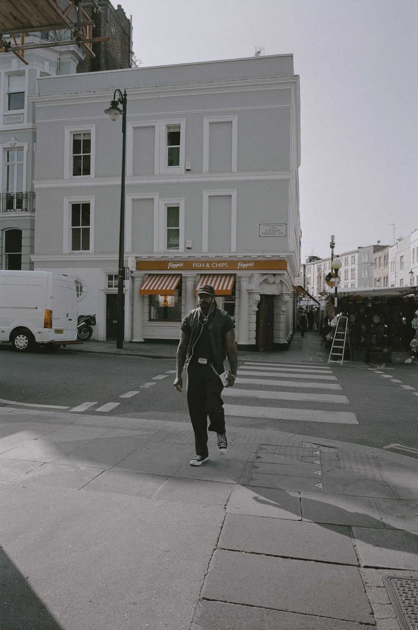 Un uomo attraversa un incrocio pedonale in una strada cittadina, con un edificio bianco sullo sfondo che ha un negozio di pesce e chips con tendine arancioni e bianche a righe.