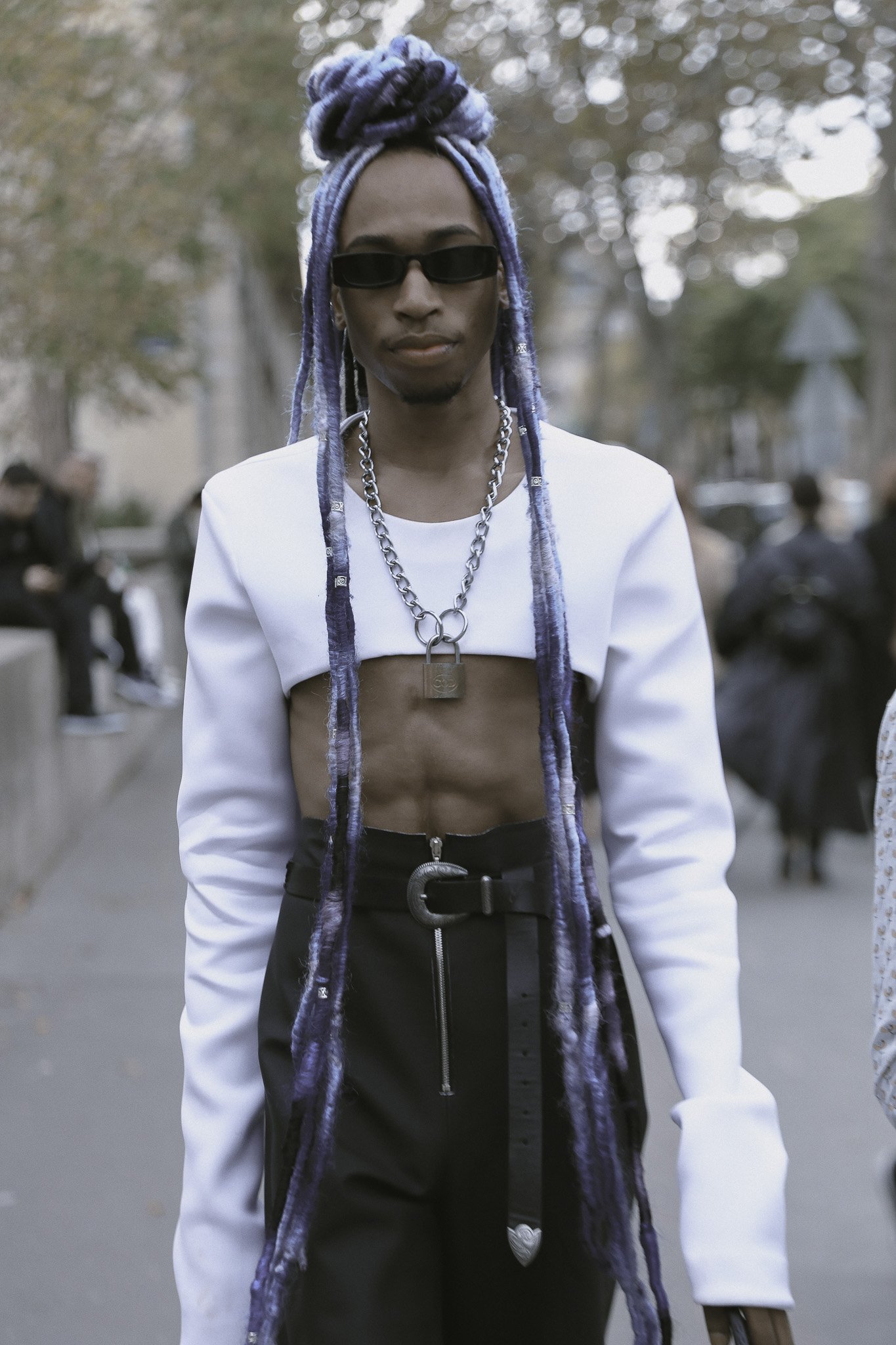 Persona con dreadlocks blu e viola, indossa occhiali da sole neri, una giacca bianca, pantaloni neri con cintura larga e chiusura con zip, e accessori come collana con catenina e lucchetto e cintura decorativa, in un contesto urbano all'aperto.