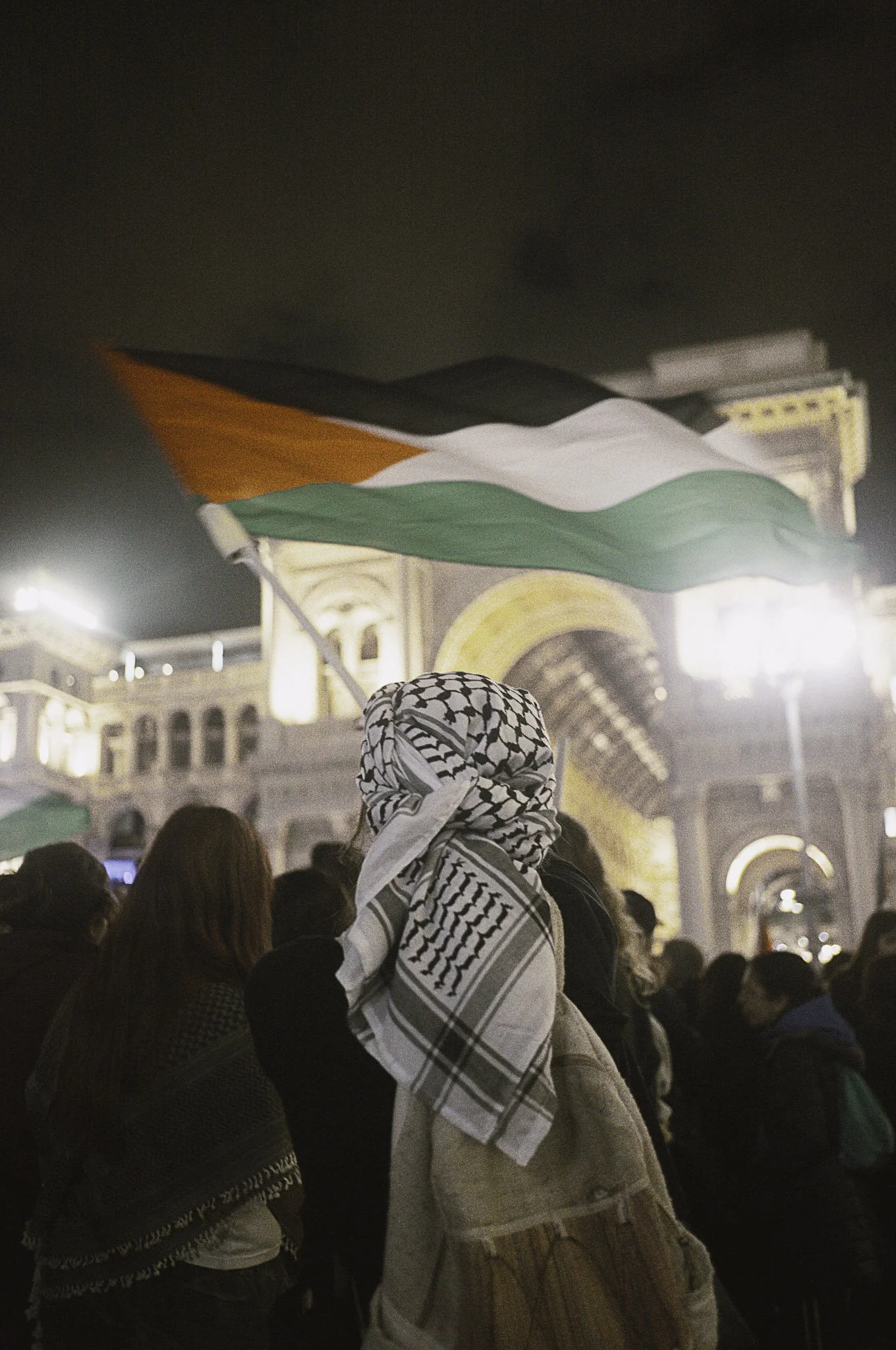 Persona con sciarpa keffiyeh e bandiera palestinese in manifestazione davanti a un edificio storico illuminato di notte.