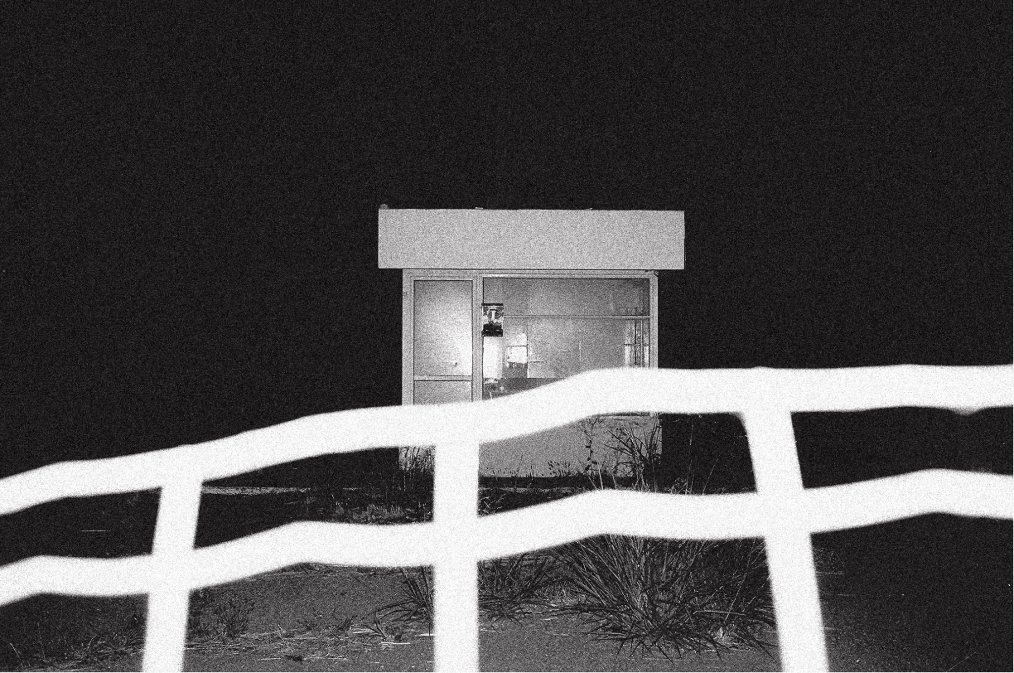 Una semplice casetta con portafinestra illuminata di notte, vista parzialmente attraverso una recinzione bianca.