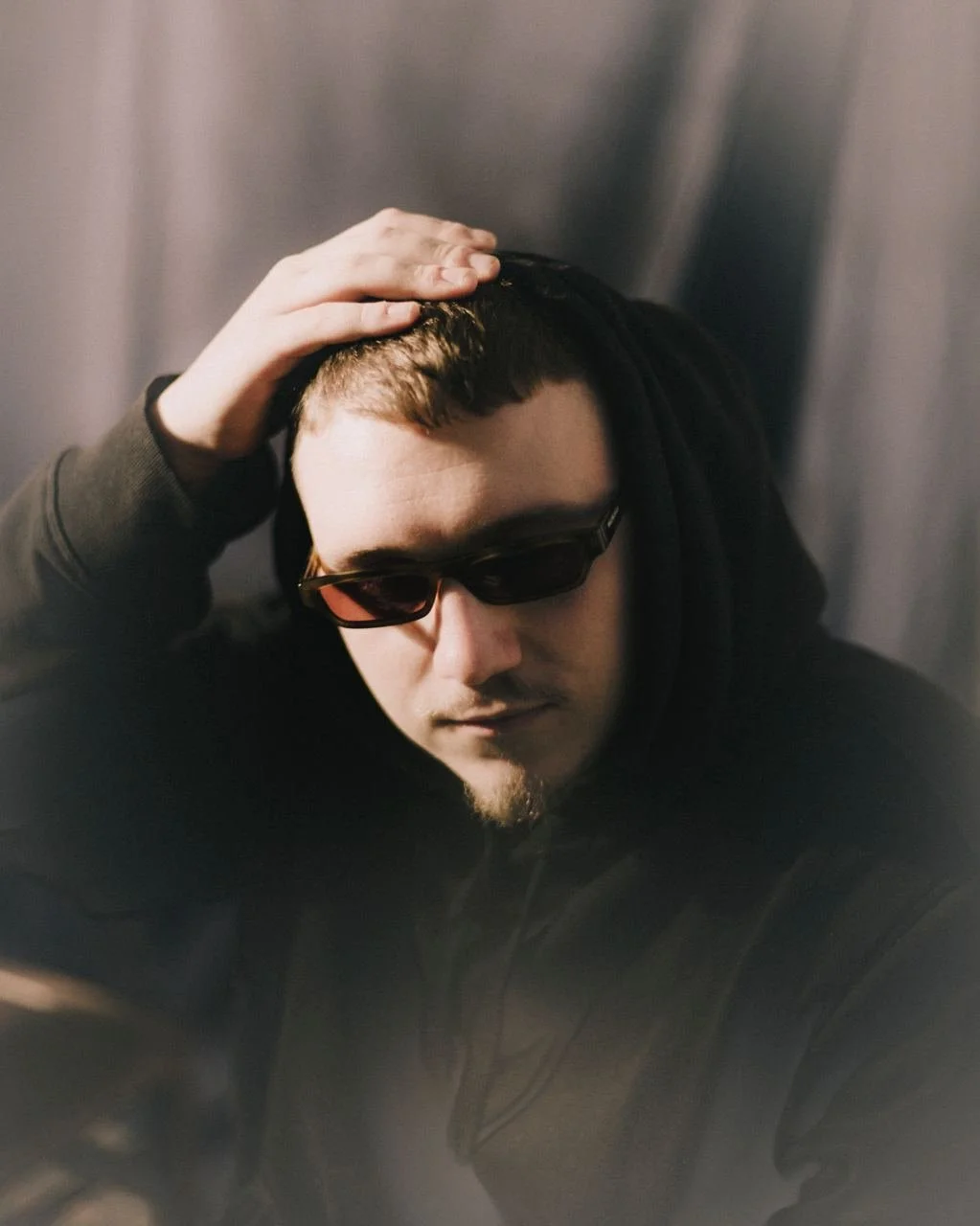 Un uomo con capelli corti e barba, indossa occhiali scuri e cappotto nero, posa con una mano sulla testa davanti a uno sfondo grigio.