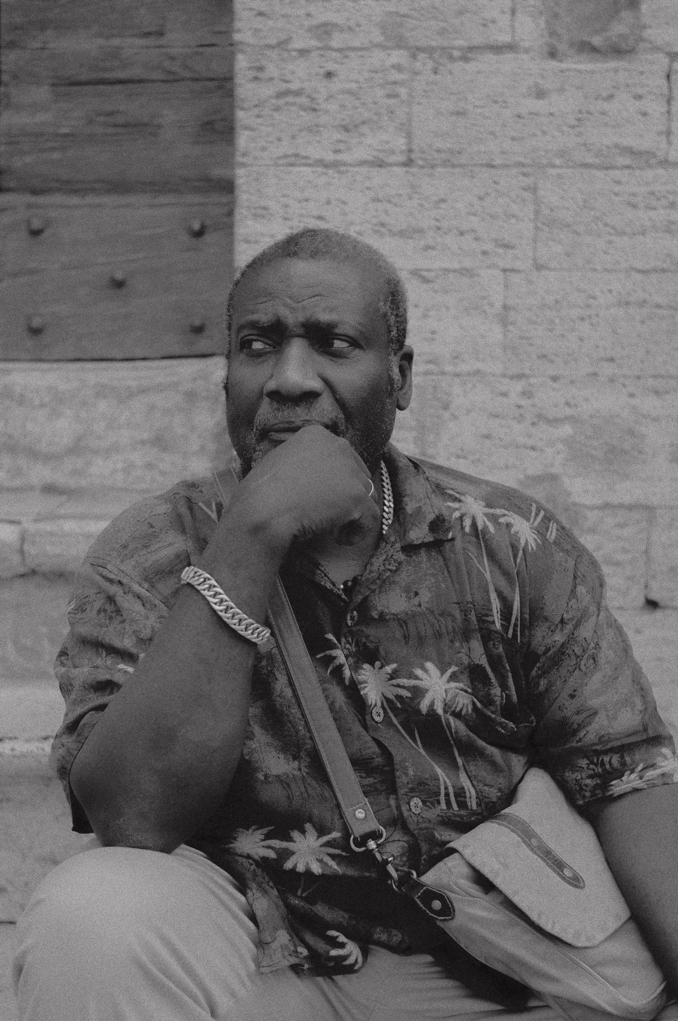 Uomo anziano con pelle scura, capelli corti grigi, vestito in camicia con motivo di palme, seduto su una sedia, appoggiando il mento sulla mano, con uno sfondo di parete in pietra