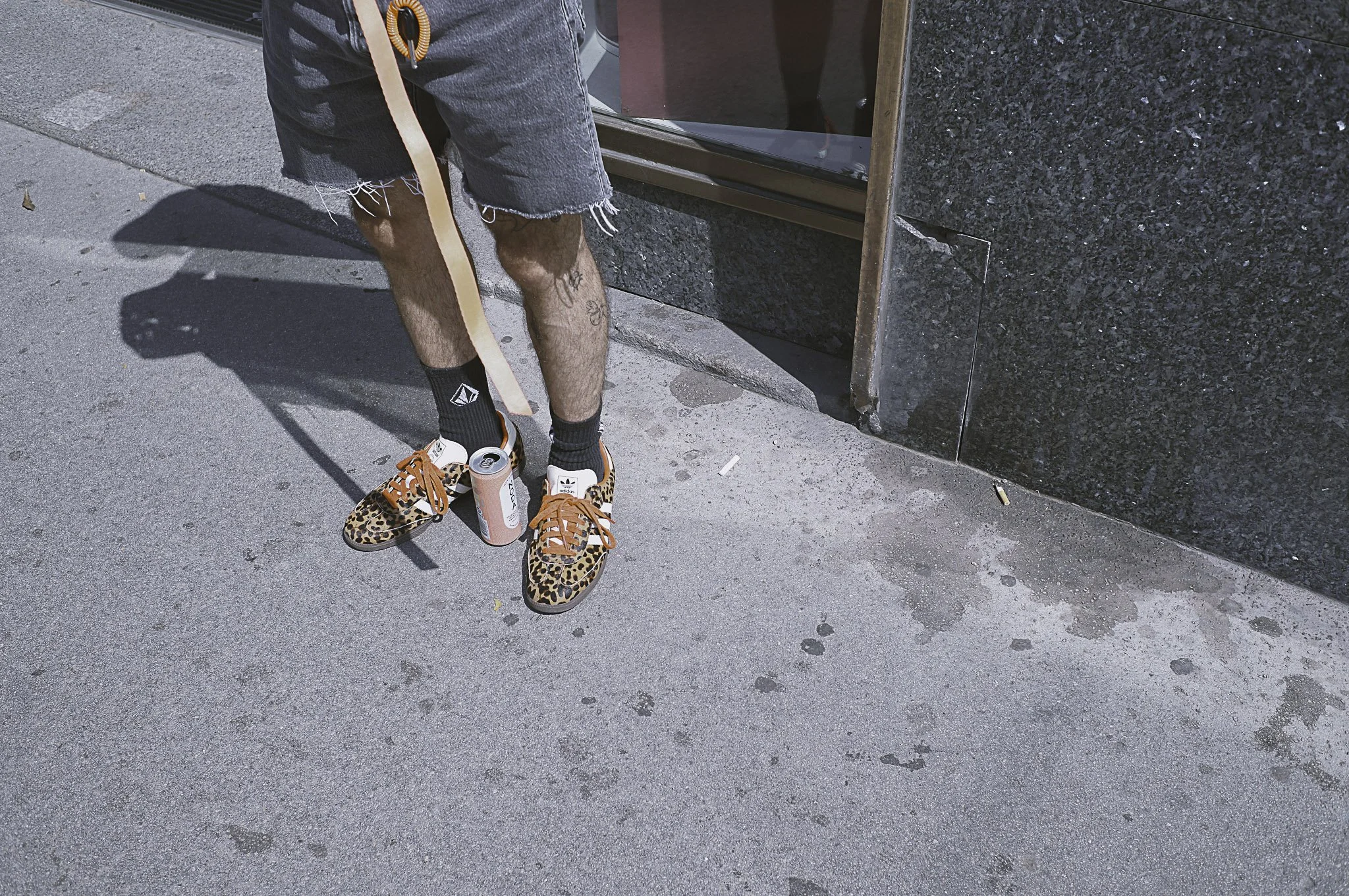 Parte inferiore di una persona con pantaloncini corti, calzini neri e scarpe con stampa leopardata, che tiene in mano una lattina e si trova davanti a una scala di cemento e un muro di pietra
