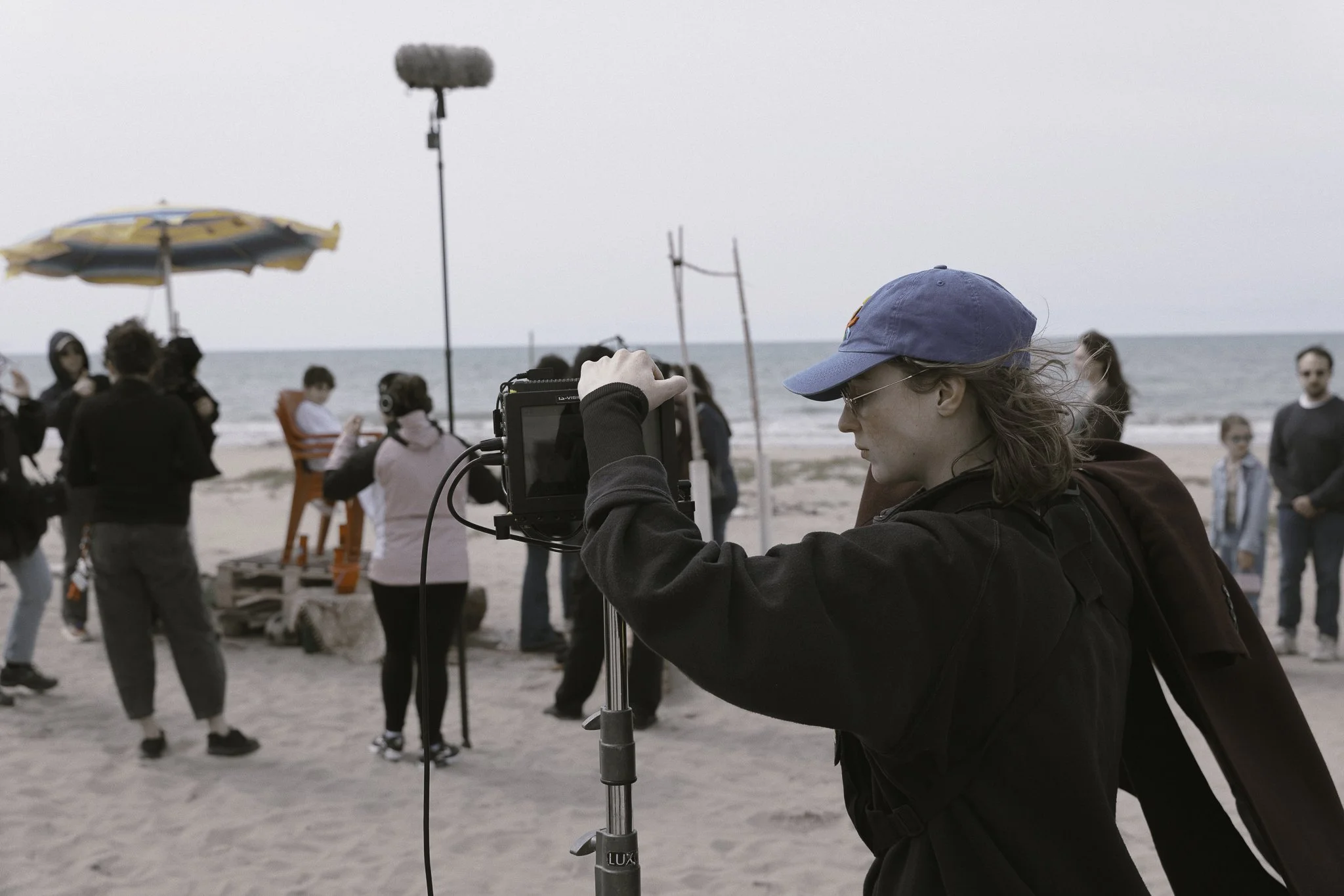 Una persona con cappellino blu e giacca nera che lavora con una videocamera in una spiaggia mentre intorno ci sono altre persone e l'oceano sullo sfondo.