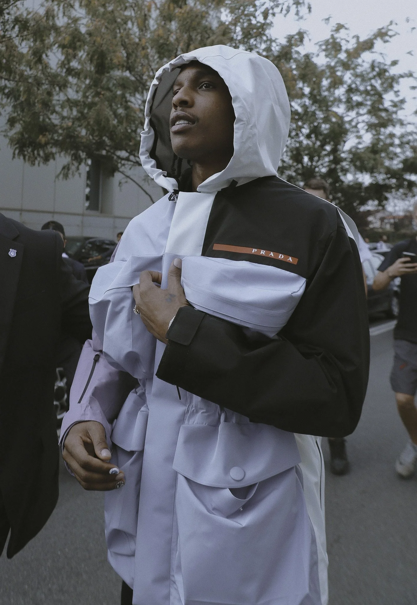 Uomo con cappotto irreale con cappuccio che indossa un giubbotto Prada, che si tiene il cuore con una mano mentre cammina.