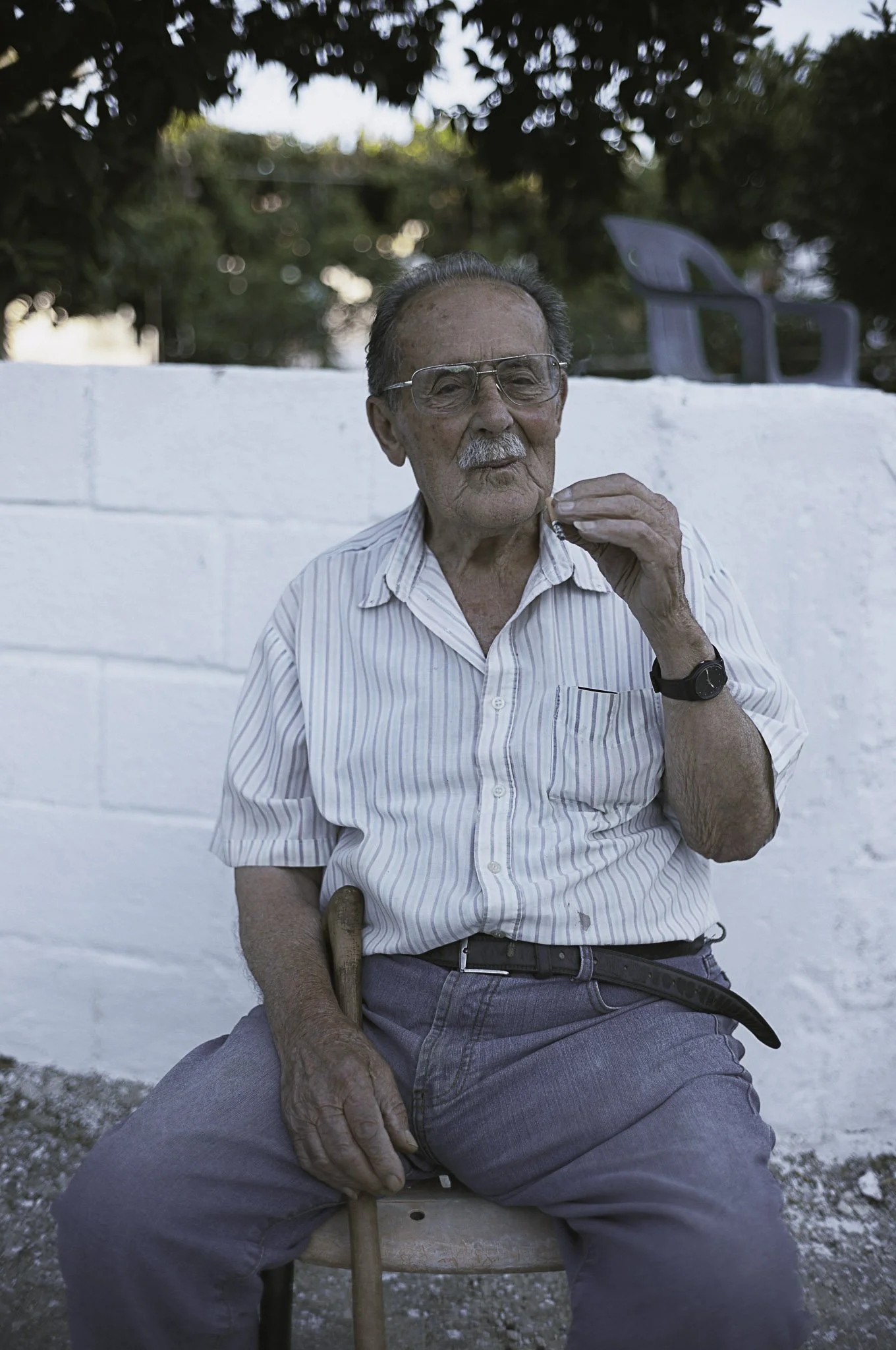 Un uomo anziano seduto su una sedia, indossa occhiali, camicia a righe e pantaloni grigi, tiene un bastone in mano e guarda con un'espressione tranquilla. Sullo sfondo, una parete bianca e alcune sedie di plastica.