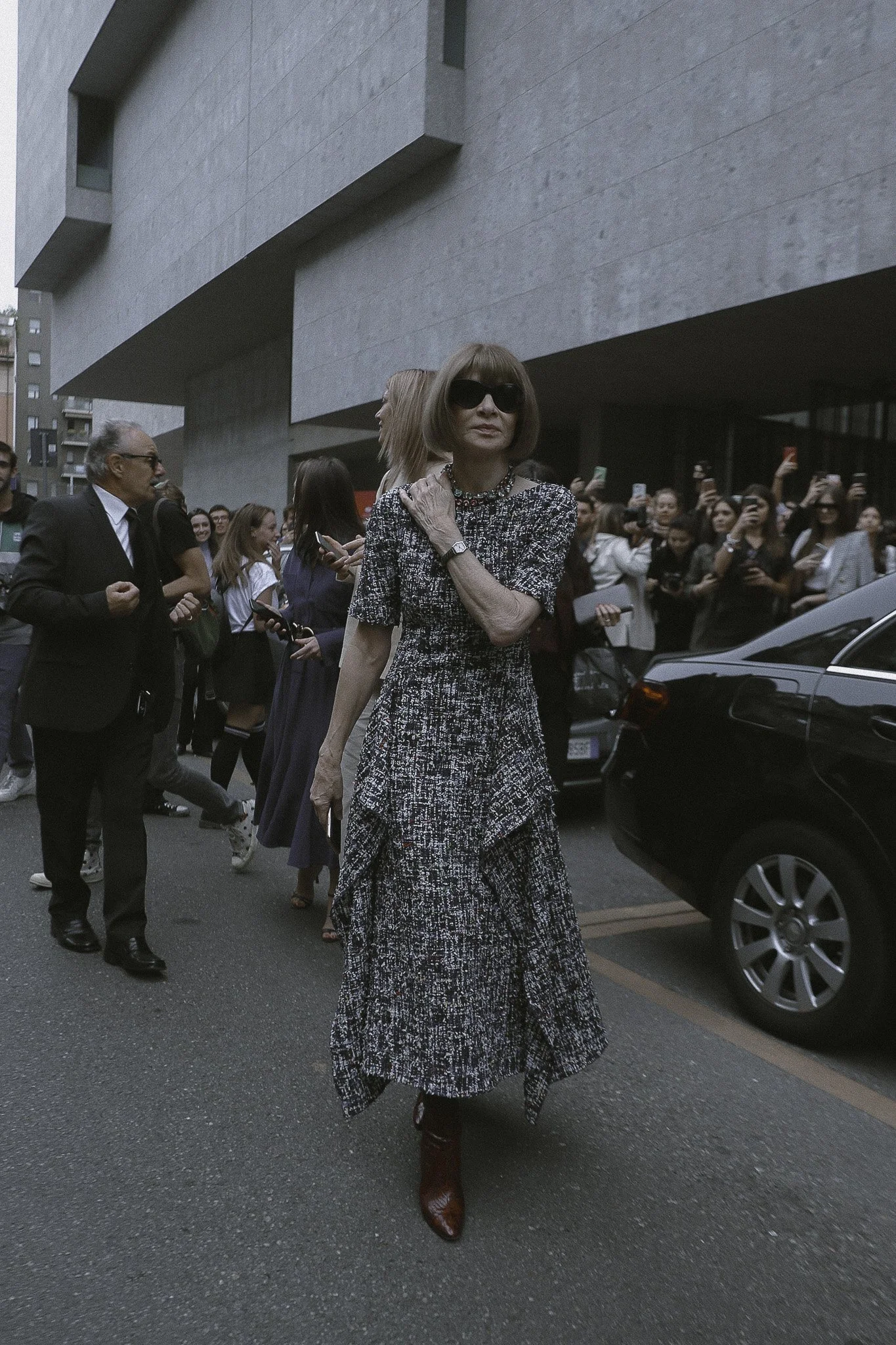 Una donna con occhiali scuri indossa un abito lungo a maglia e stivaletti marroni, cammina tra una folla di persone che la stanno filmando e fotografando fuori da un edificio moderno.
