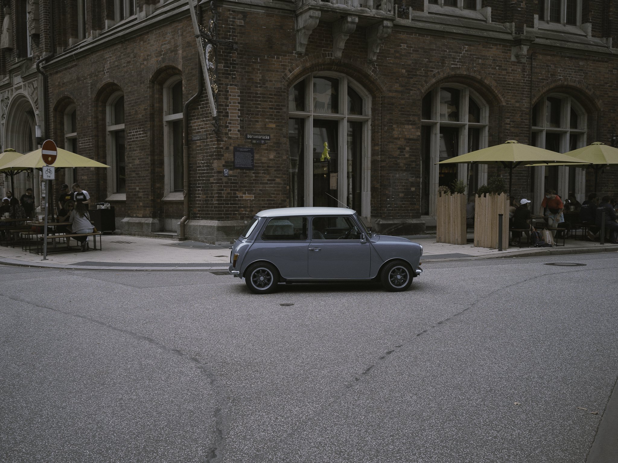 Una piccola macchina Classica di colore grigio parcheggiata su strada accanto a un edificio in mattoni con grandi finestre, patio con tavoli con ombrelloni gialli e persone sedute e mangiando.