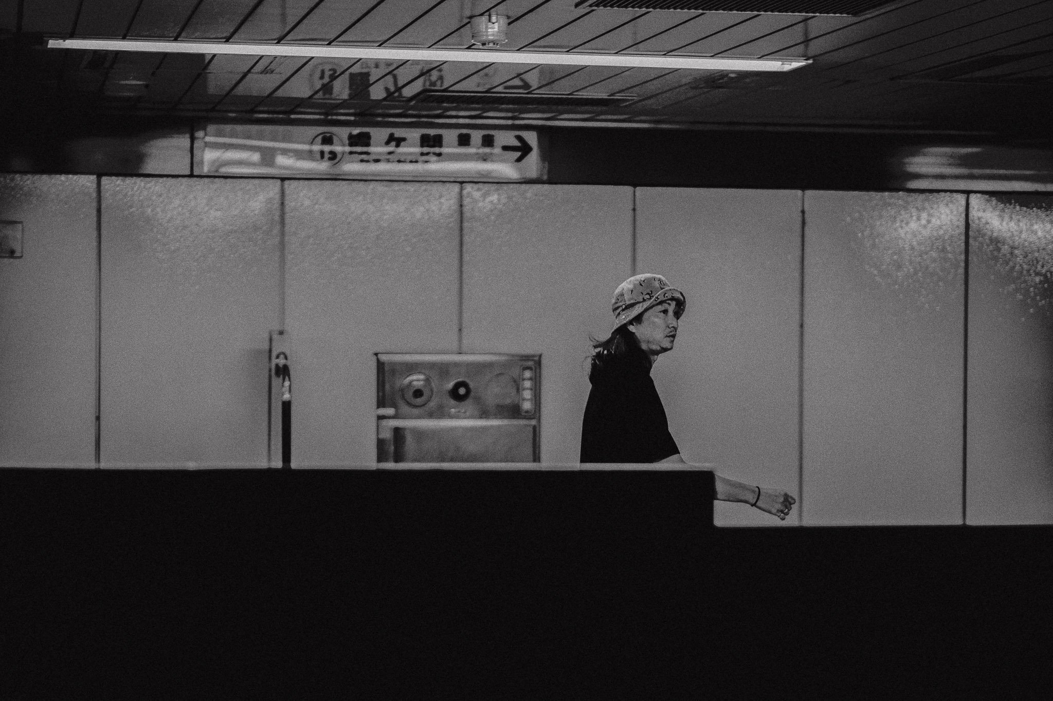 Donna con cappello che cammina in un ambiente interno, probabilmente una stazione o un edificio pubblico, in bianco e nero.